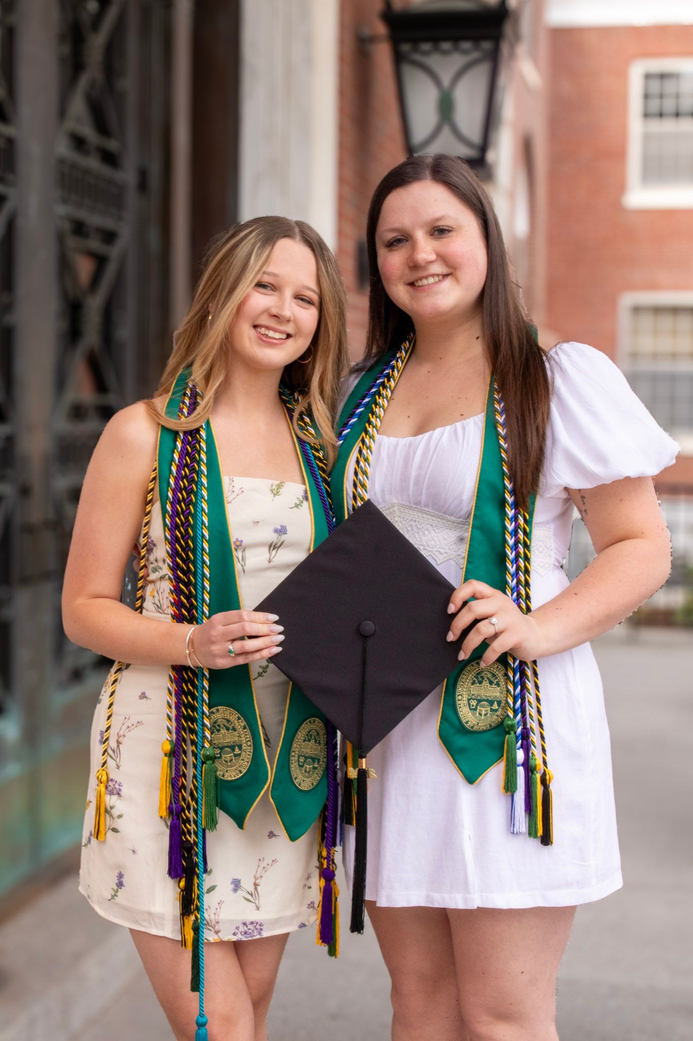 Two friends wearing graduation attire and green decorative cords pose together for a series of celebratory photos.