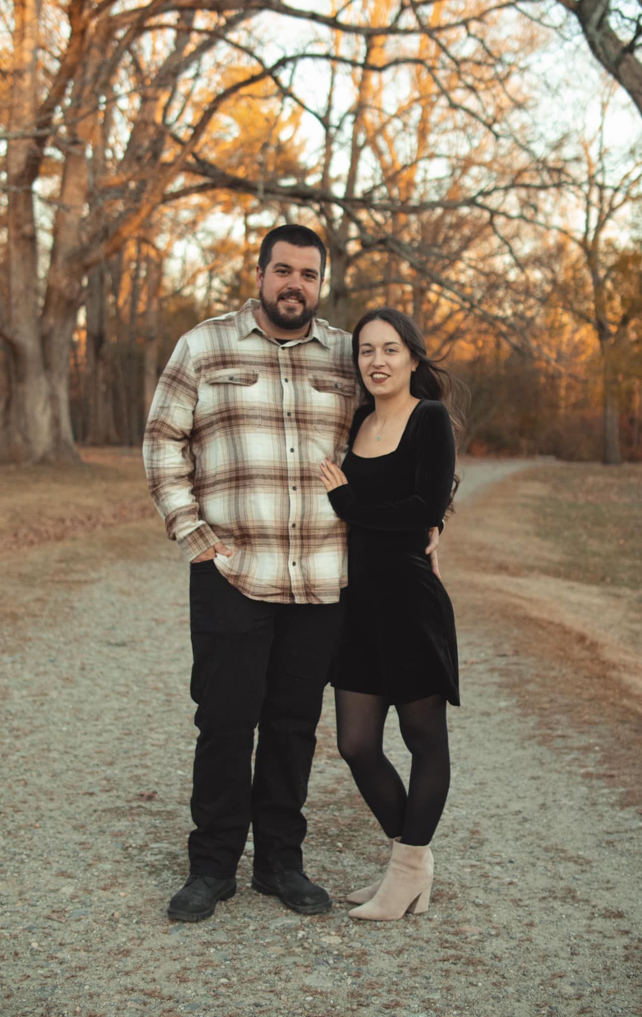 A couple poses together on a fall day in a park setting with autumn trees in the background.