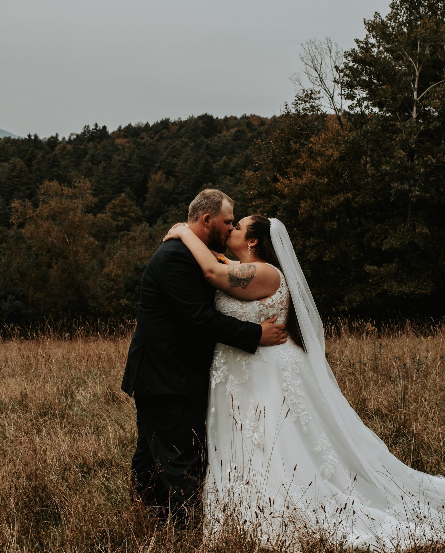 A romantic wedding couple embraces in a golden autumn field with trees in the background.