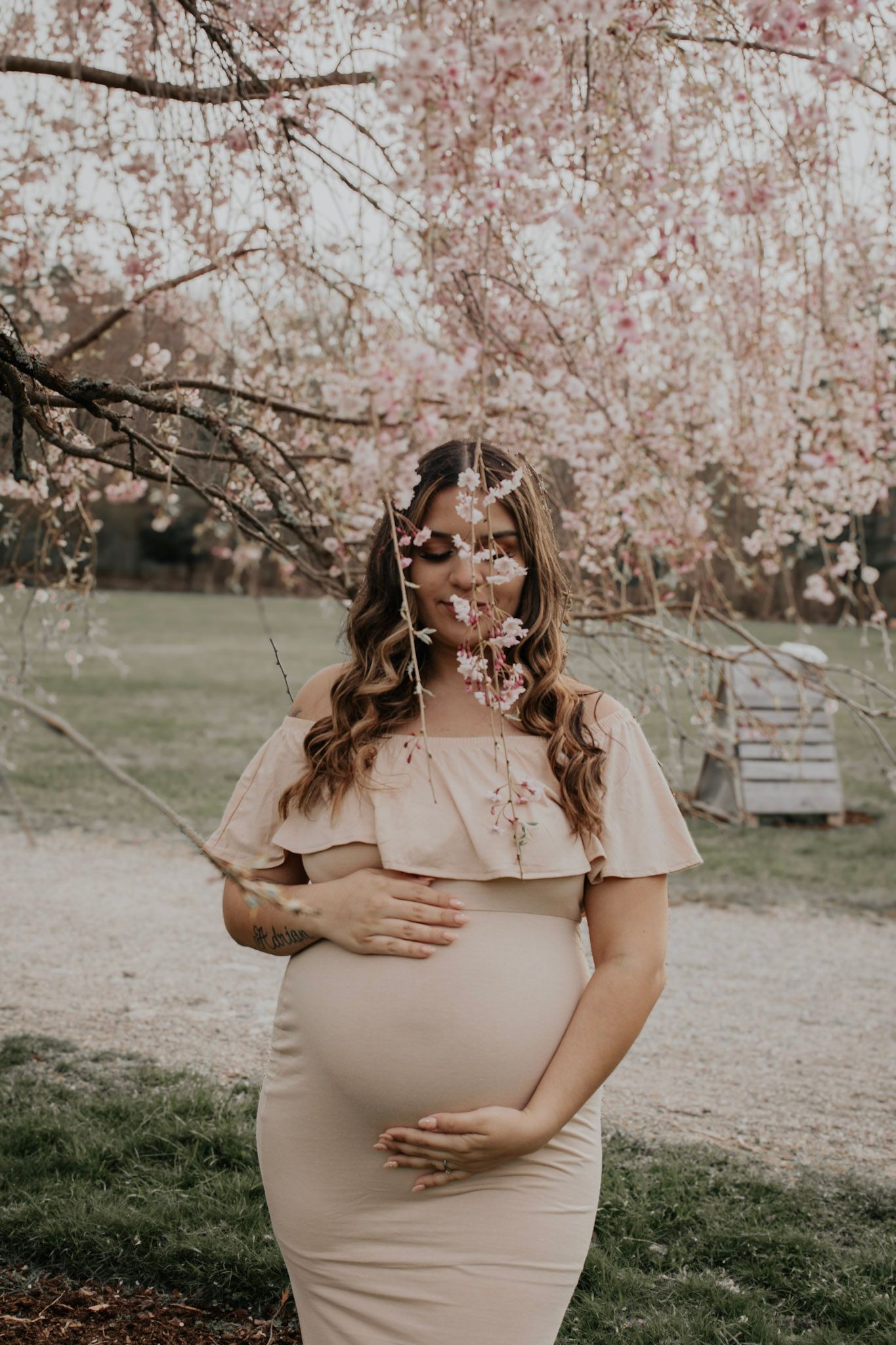 A pregnant woman in a beige dress stands under blooming cherry blossom trees during a spring maternity photoshoot.