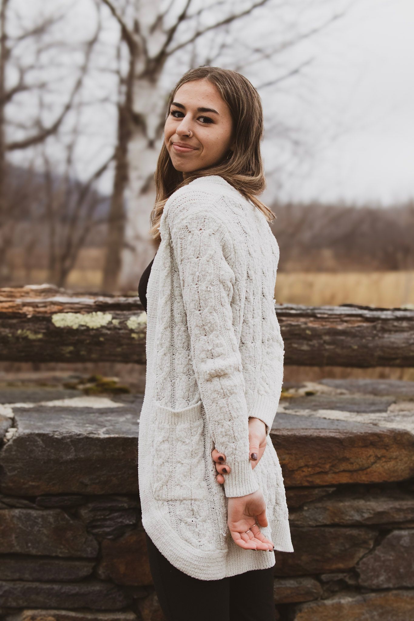 A person in a cozy white cable knit sweater poses against a rustic stone wall backdrop on an autumn day.