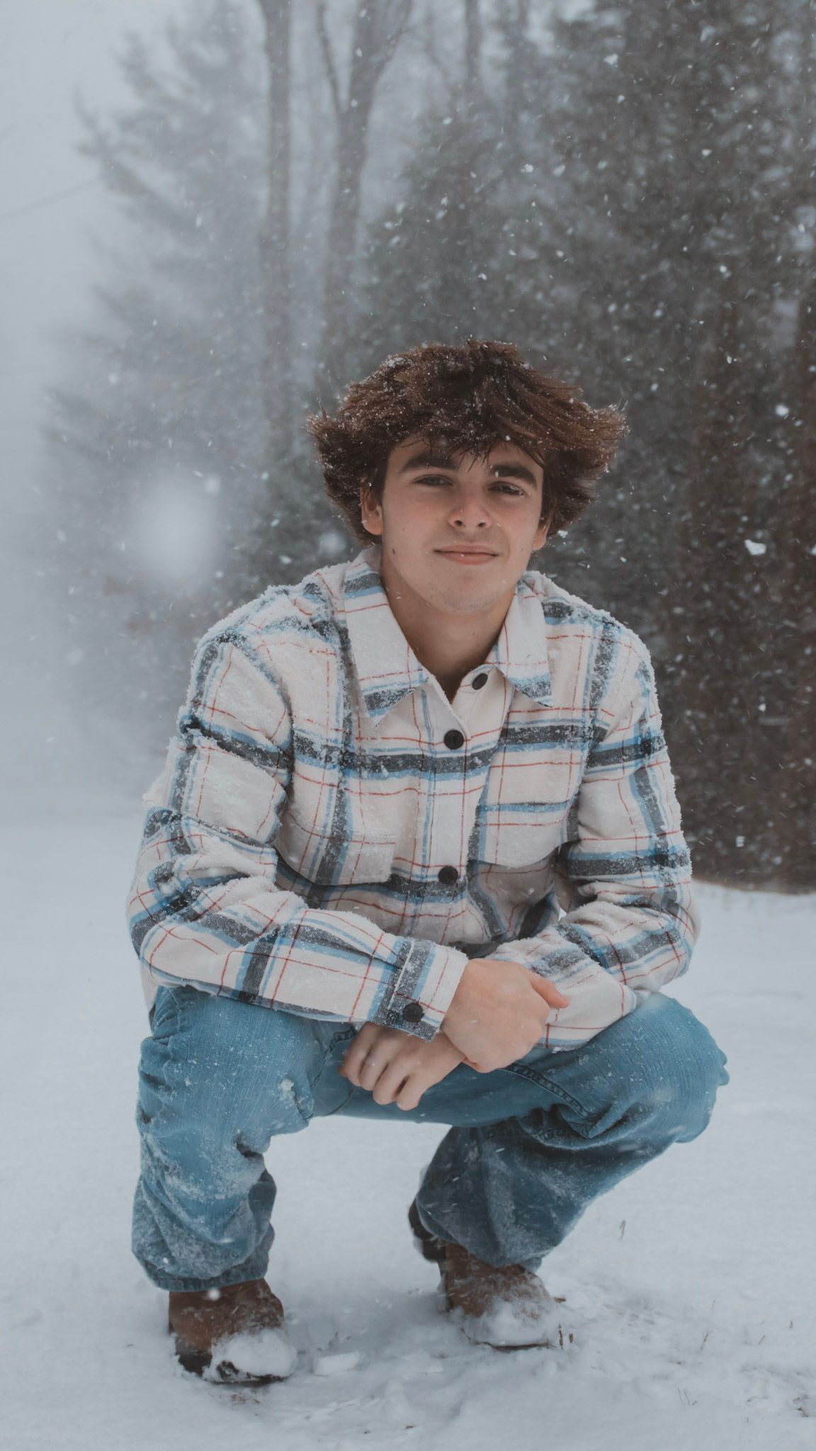 A person in a plaid shirt and jeans crouches in the snow while snowflakes fall in a misty winter forest setting.