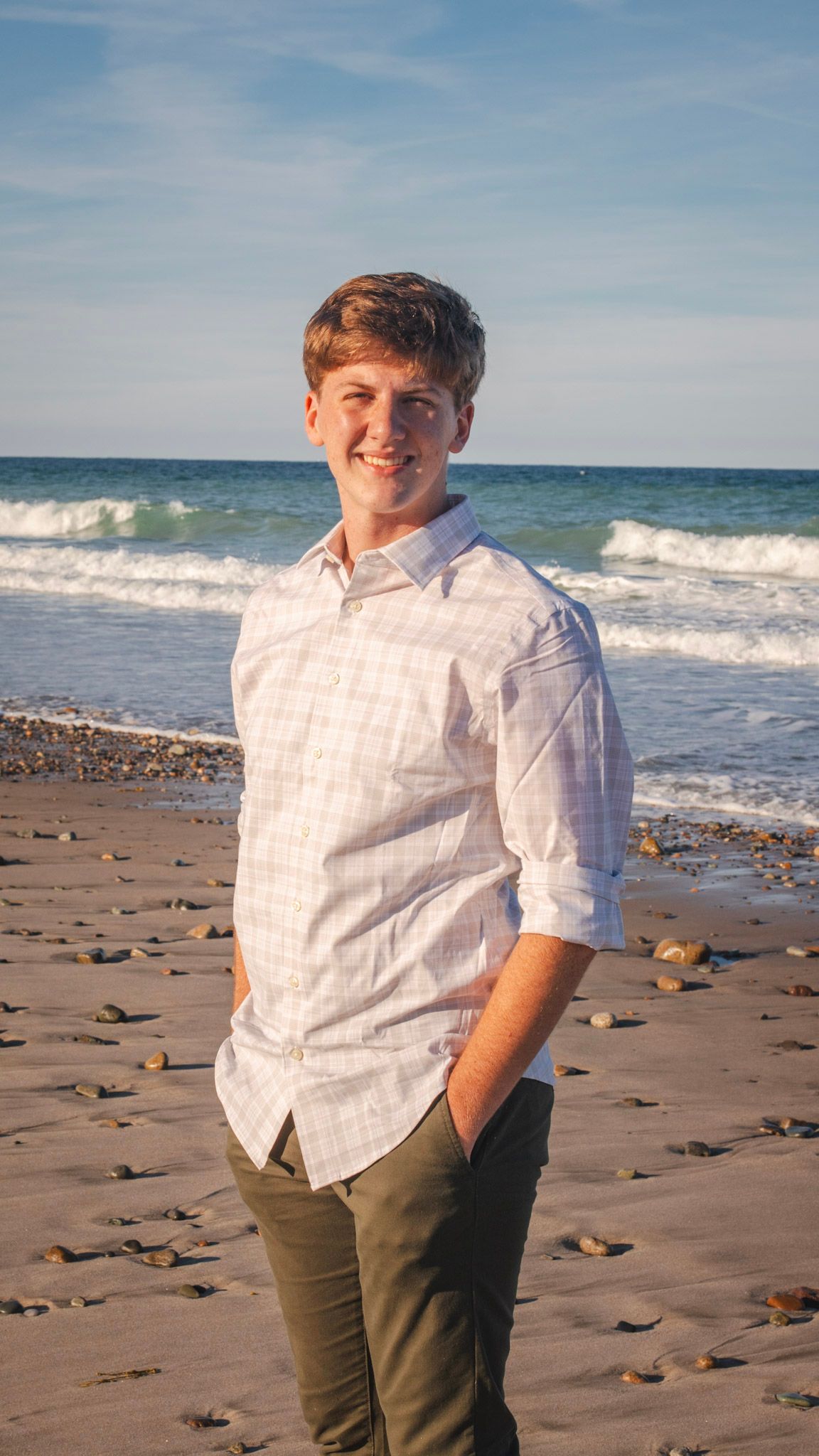 A person in a white button-down shirt and olive pants poses on a rocky beach with ocean waves in the background.