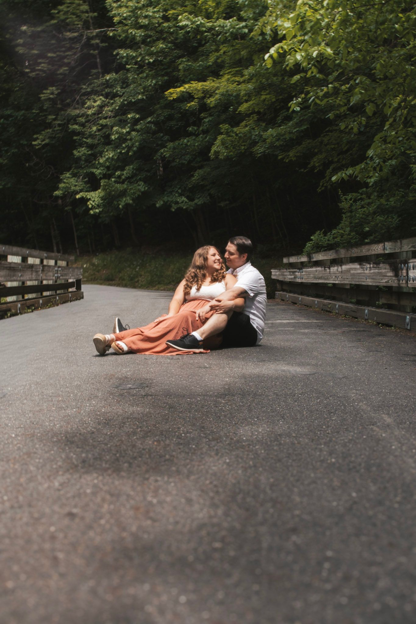 A couple sits intimately together on a paved pathway surrounded by lush green trees and wooden railings.