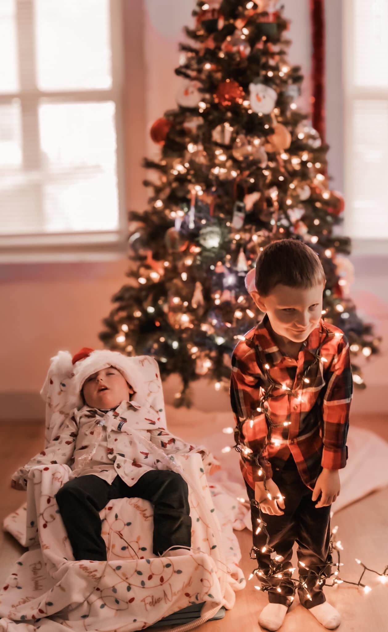 Two children playing with Christmas lights in front of decorated tree.