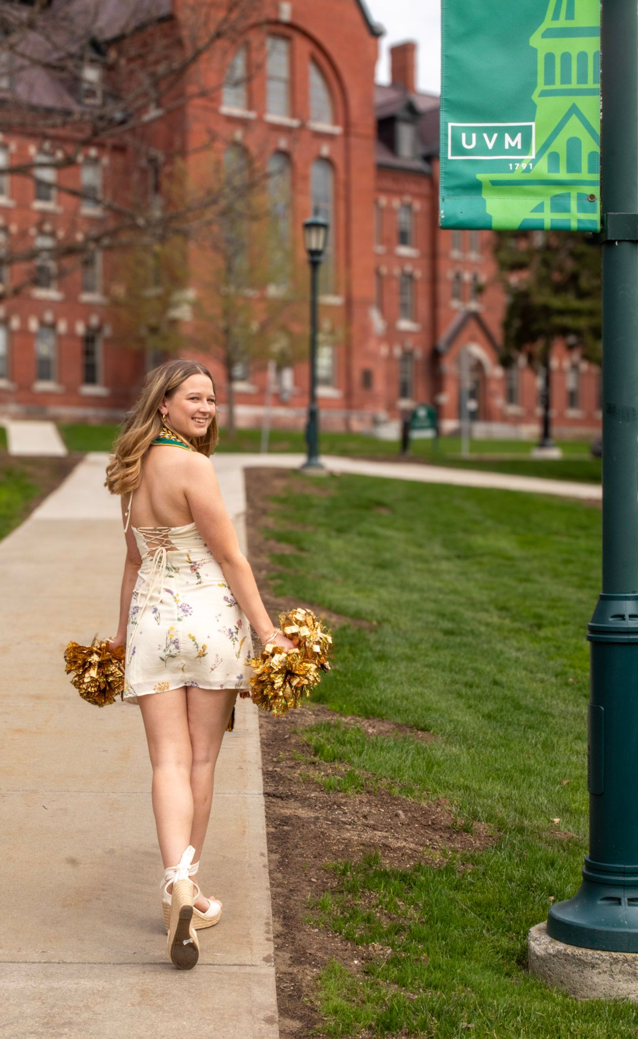 A cheerleader in a white dress and gold pom poms poses near historic brick buildings and green lampposts on a college campus.