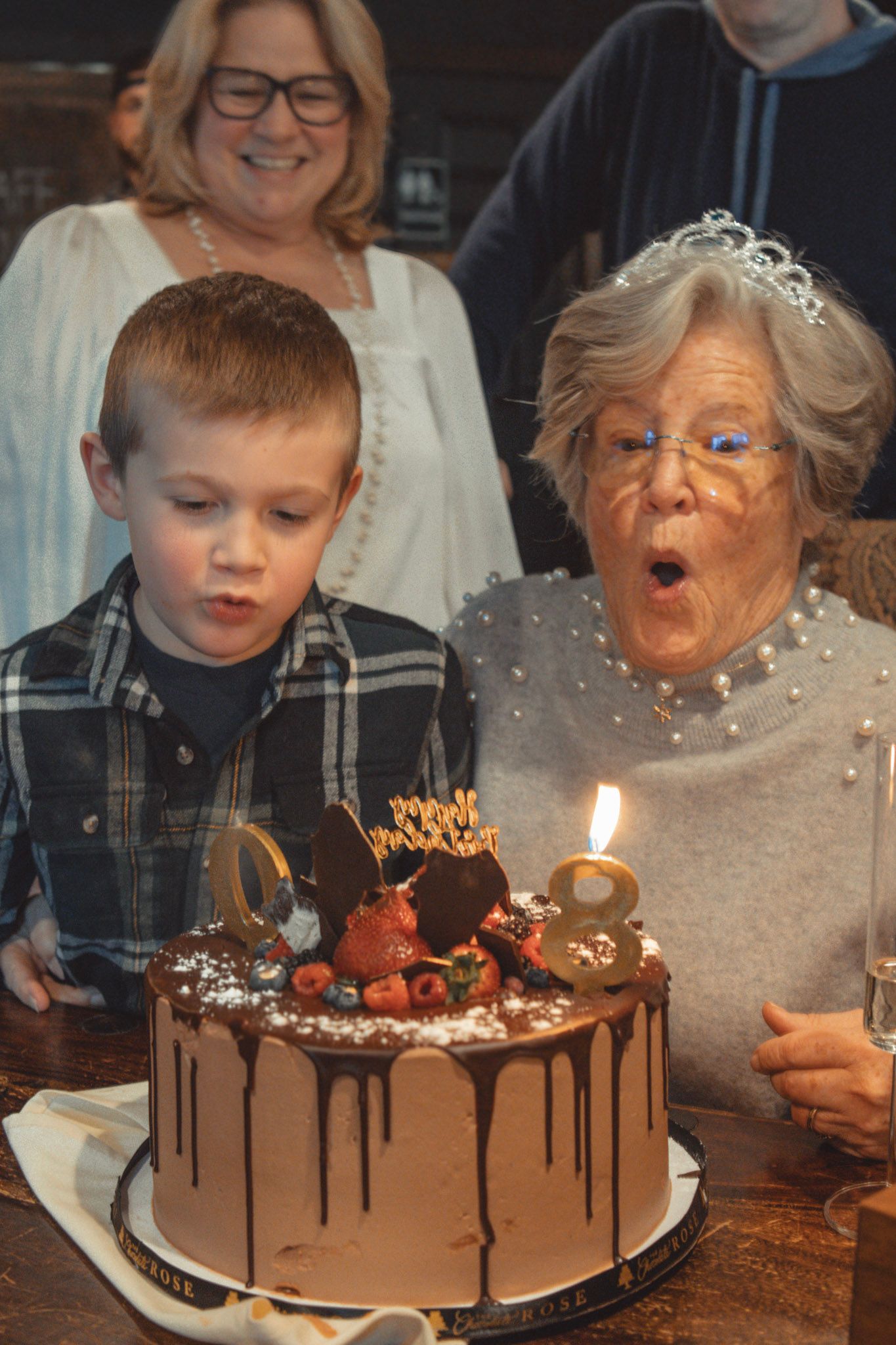 A chocolate birthday cake with dripping ganache and lit candles being celebrated by family members.