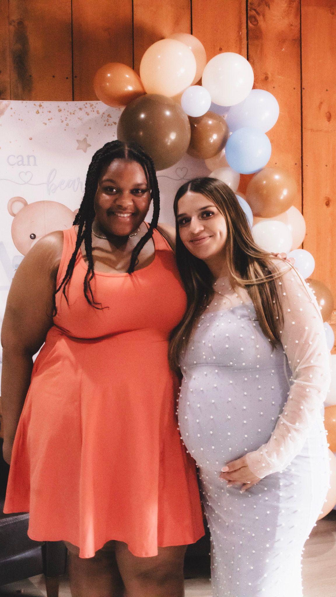 Two friends pose together at a celebration with brown and white balloons in the background against a wooden wall.