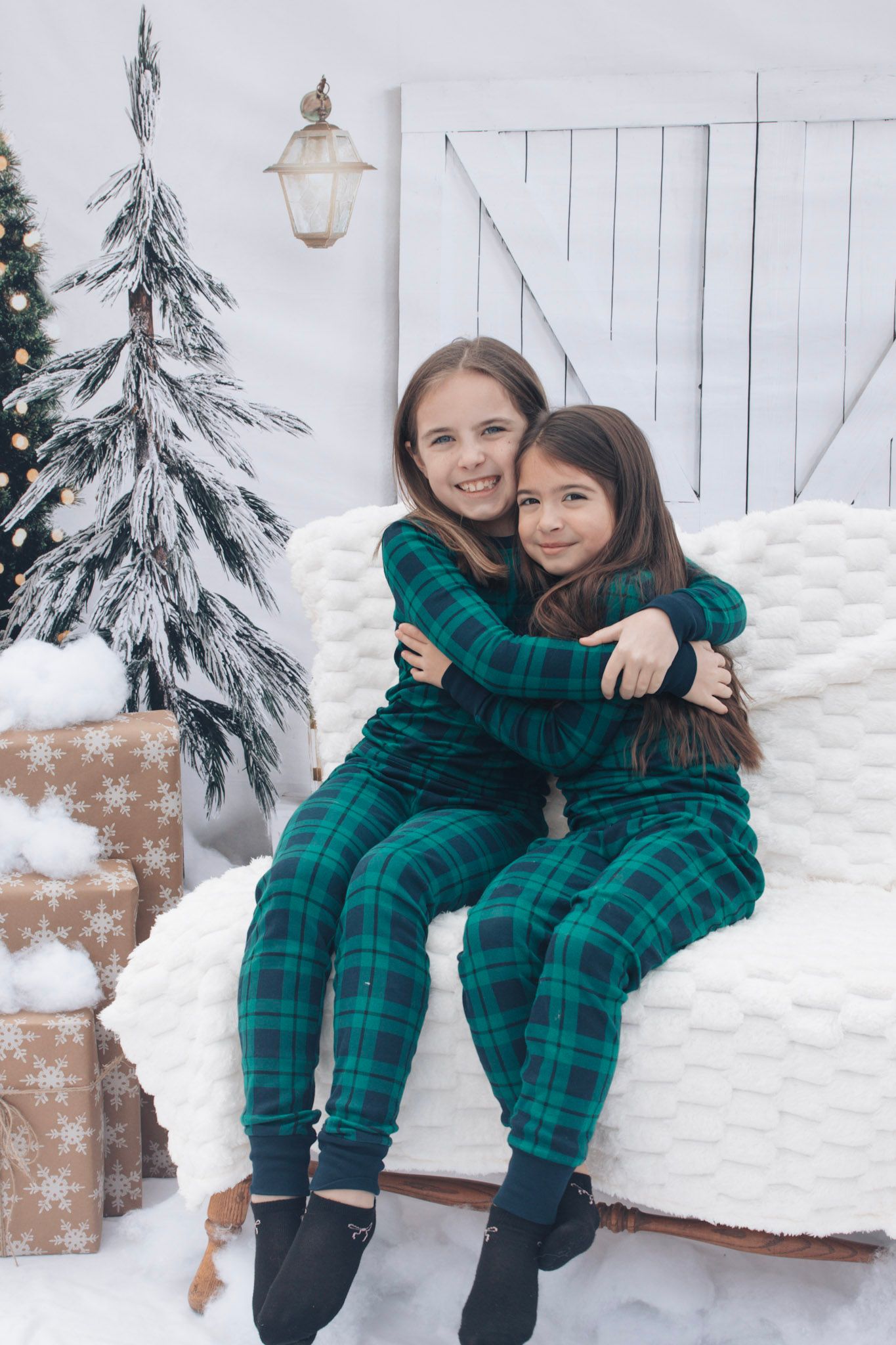 Two people in matching green plaid pajamas sitting together in a snowy winter scene with evergreen trees.