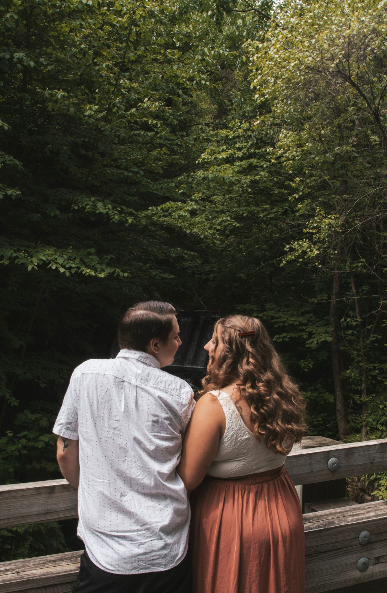 A couple sits together on a wooden bridge surrounded by lush green forest scenery.