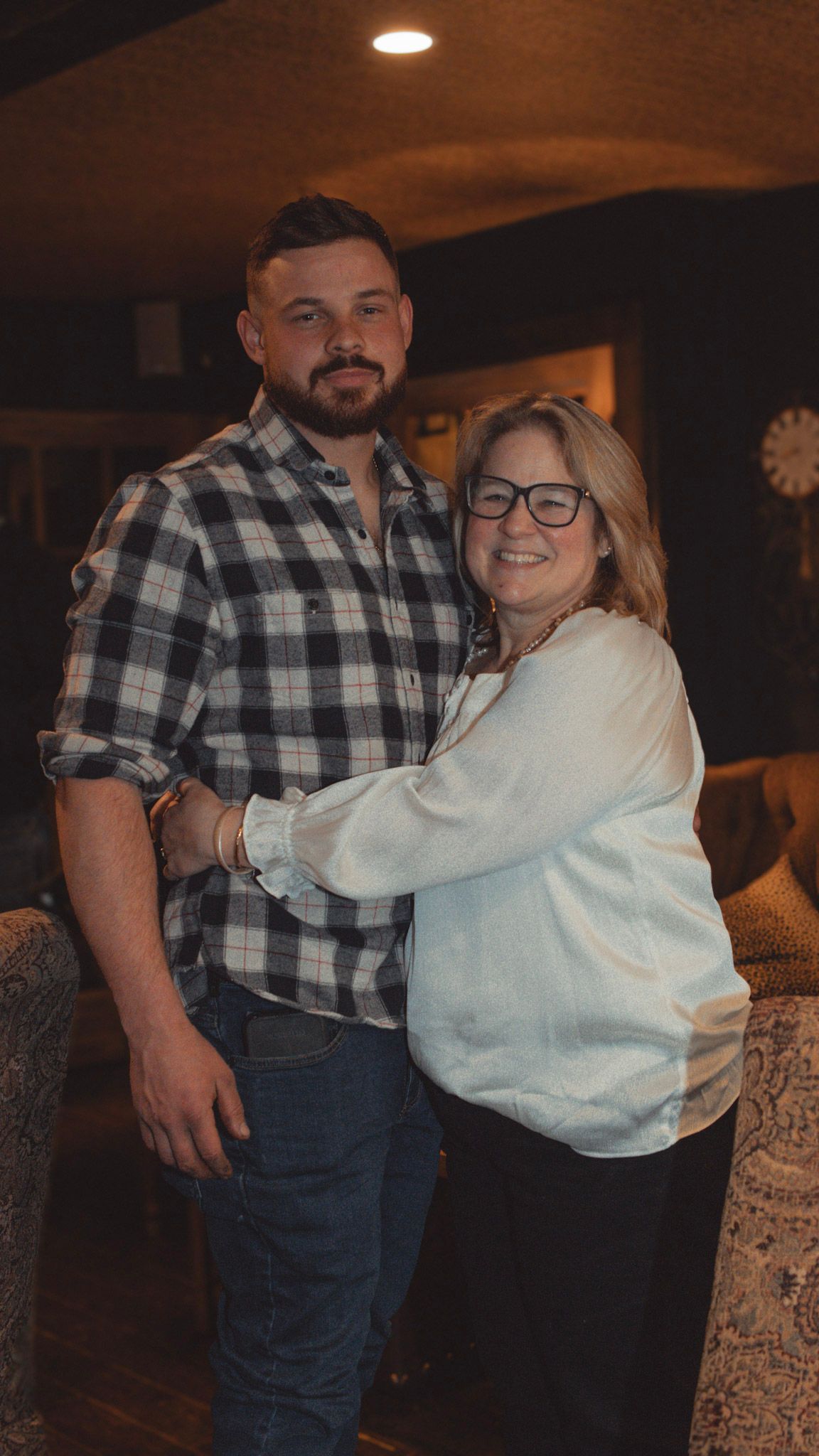 A couple embraces while dancing together in a dimly lit room, wearing casual attire including a plaid shirt and white sweater.