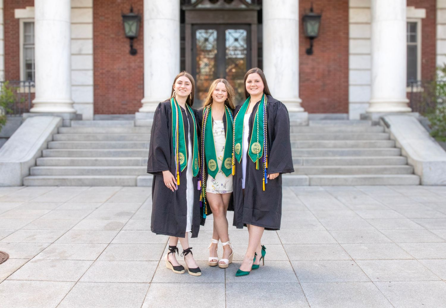 Graduates in black caps and gowns gather on stone steps outside a brick building for graduation photos.