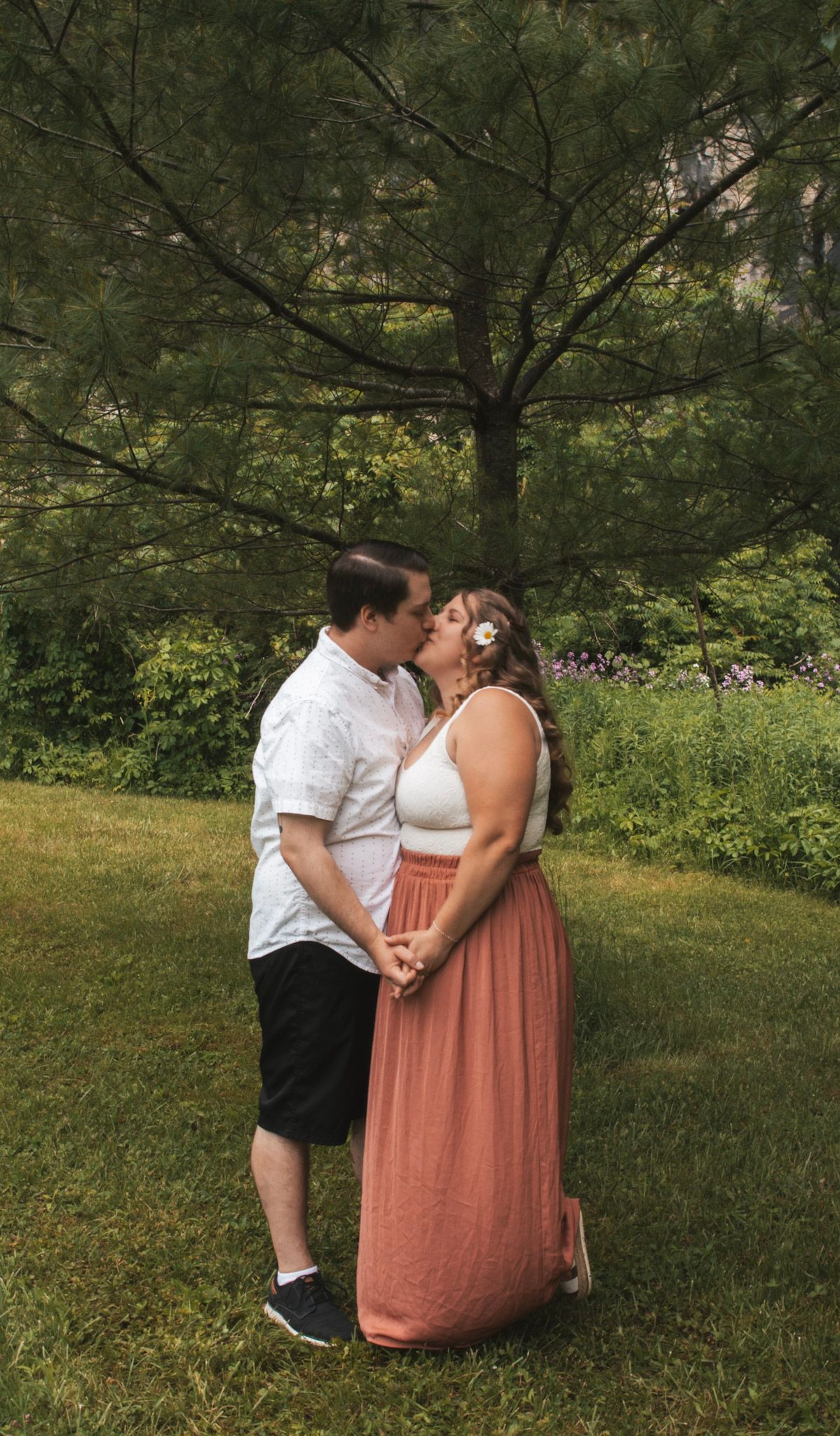 A couple shares an intimate kiss in a lush green garden setting while wearing casual summer attire.