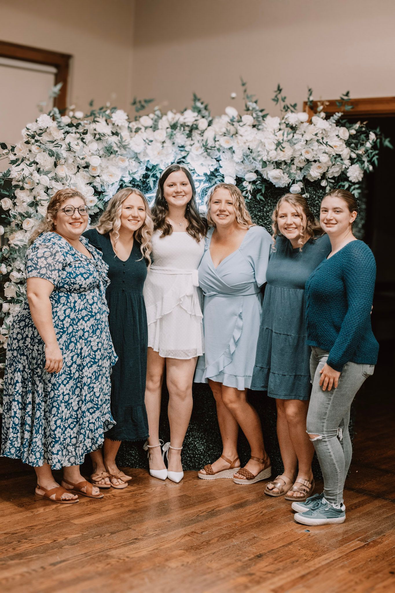 A group poses together in coordinated casual outfits against a white wall with floral decorations.