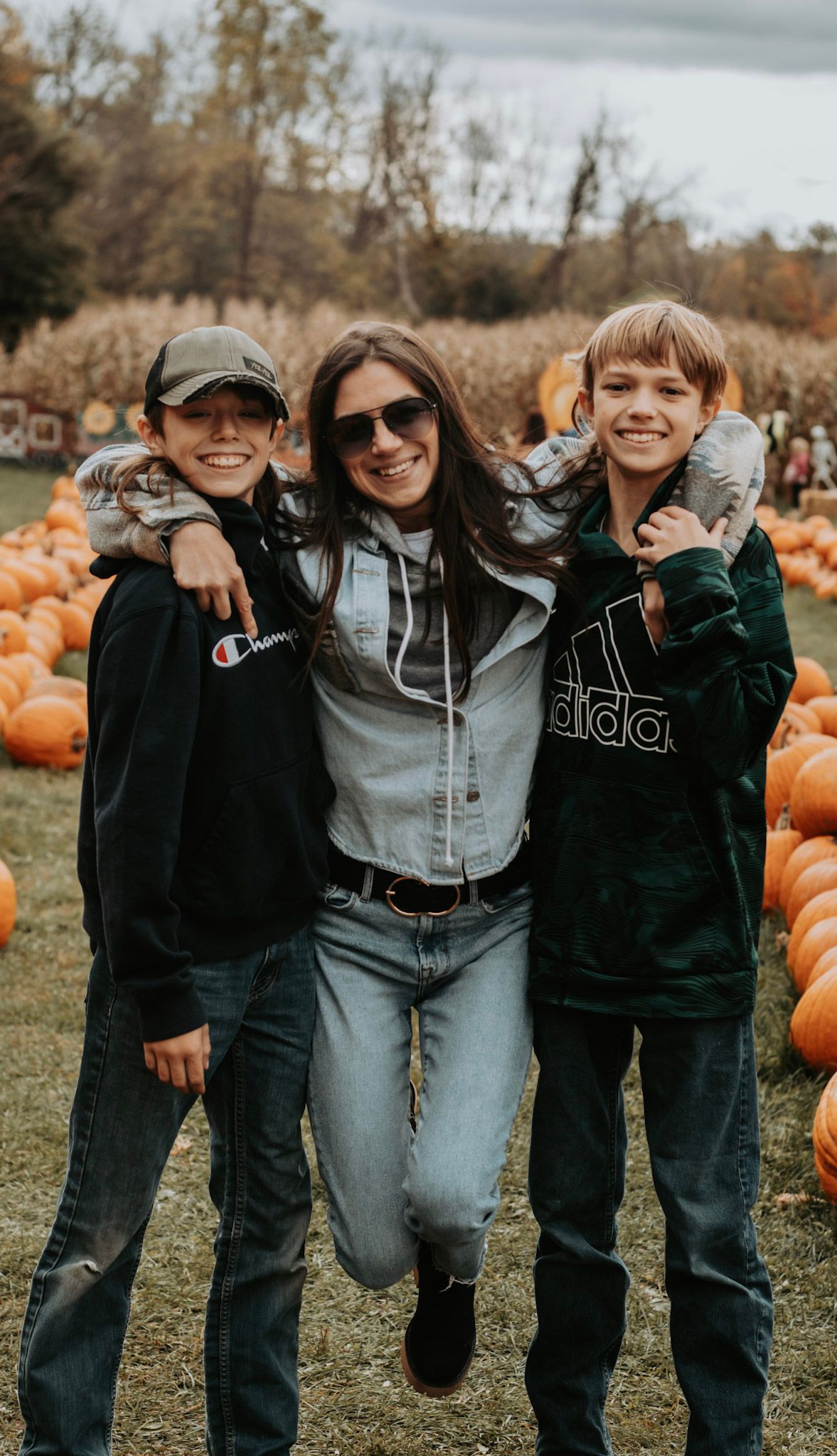 Three friends in casual outfits pose playfully together at a pumpkin patch on an autumn day.