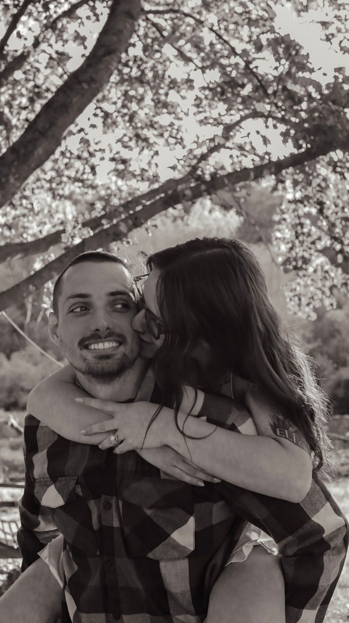 A couple embraces in a romantic piggyback pose under blooming cherry blossom trees in black and white.