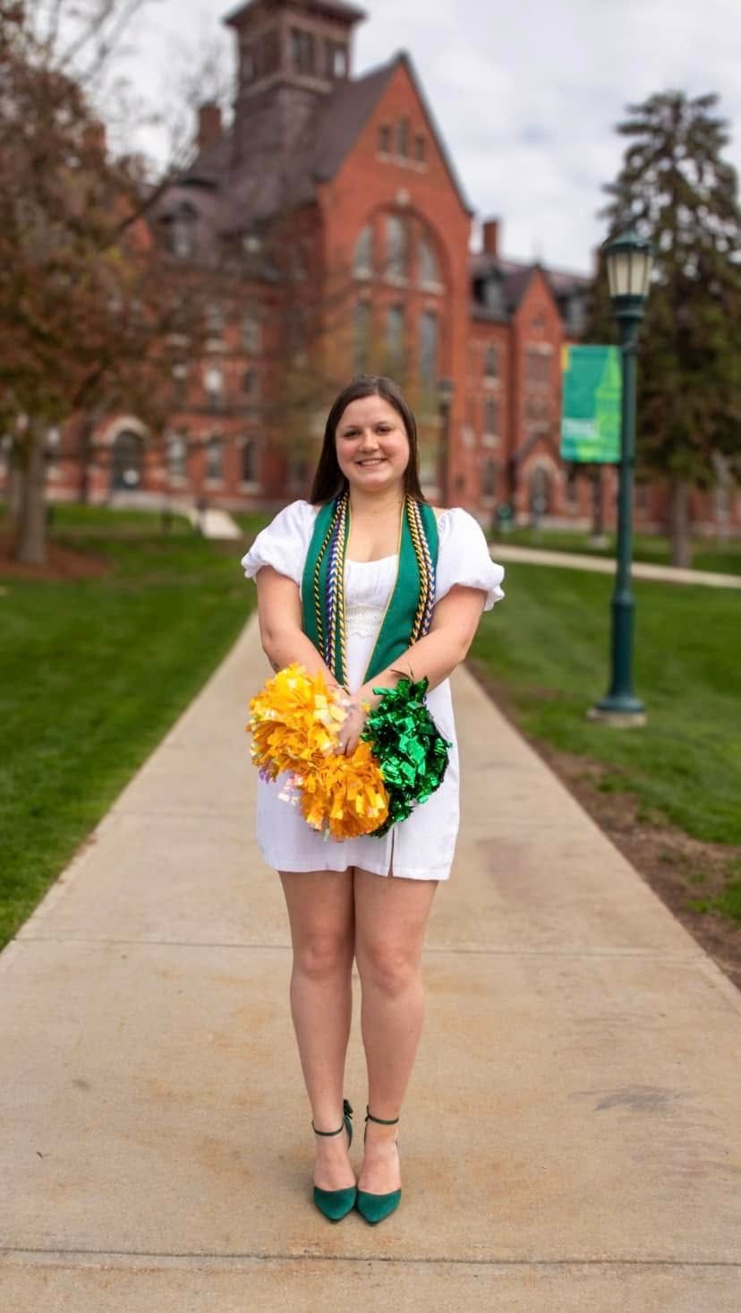 Graduate in white dress with green and gold pom poms poses on campus walkway.