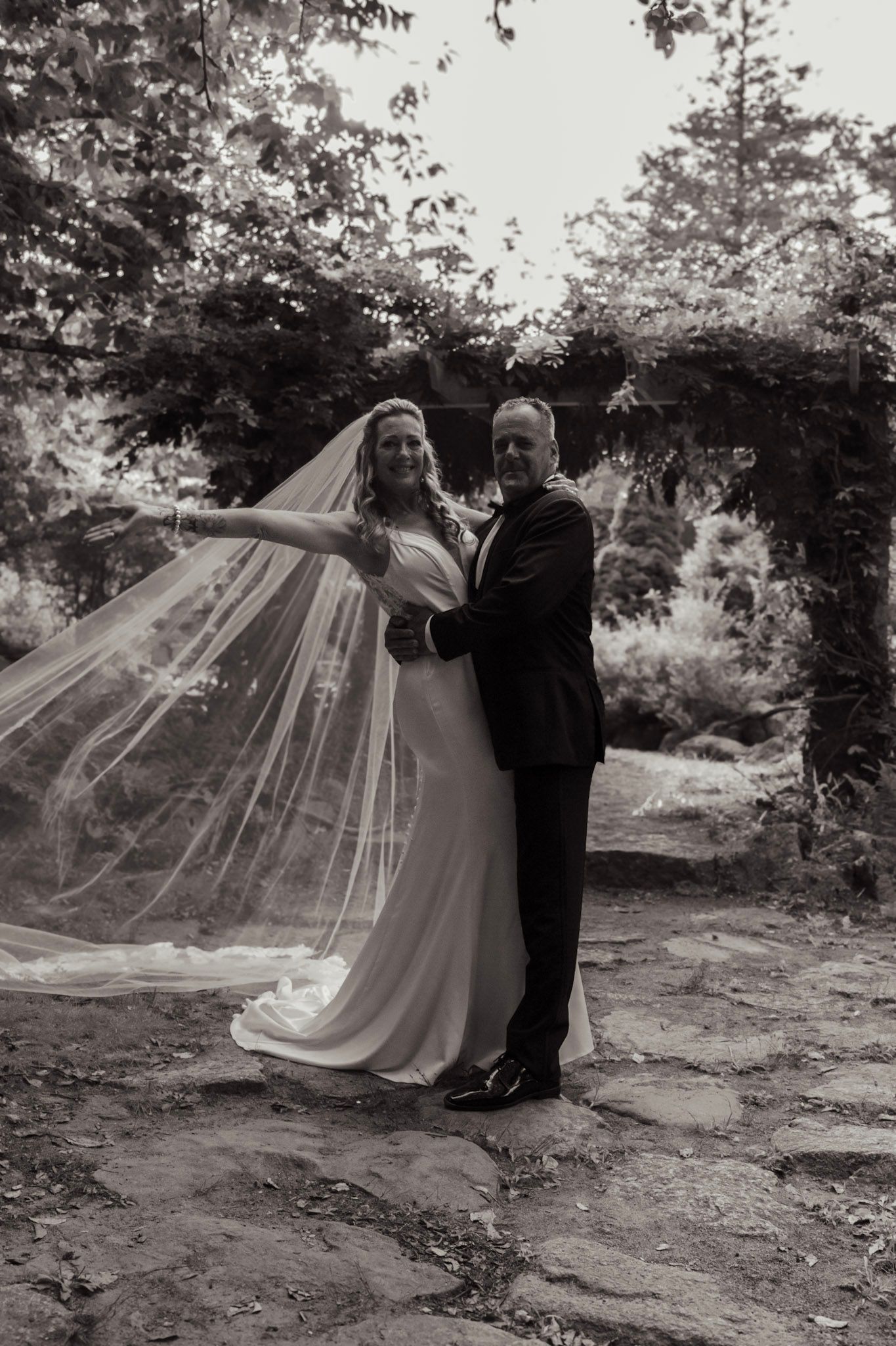 A black and white wedding portrait of a couple with a flowing veil in a garden setting.