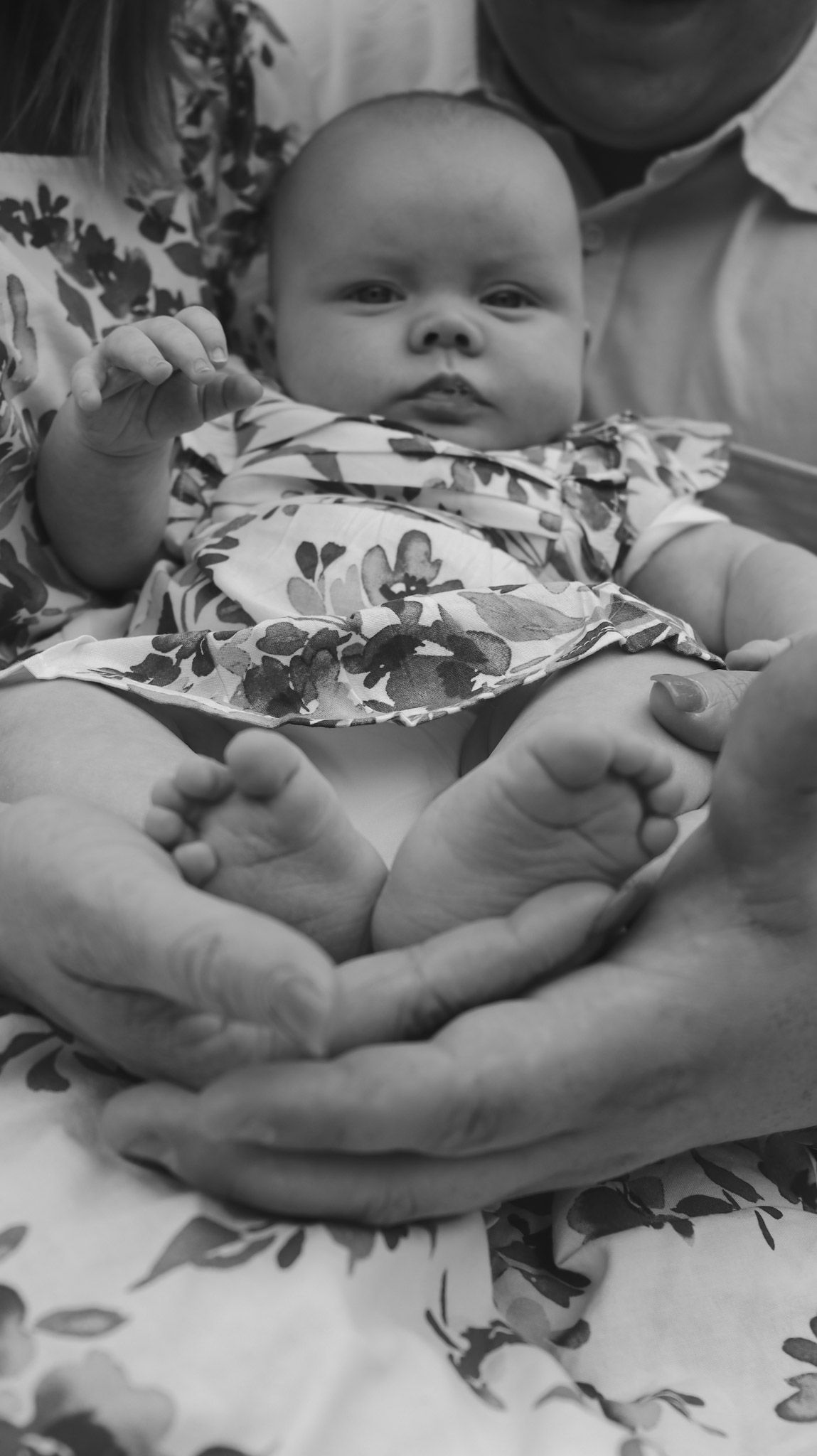 Baby wearing floral outfit rests in caring hands while looking upward in black and white photograph.