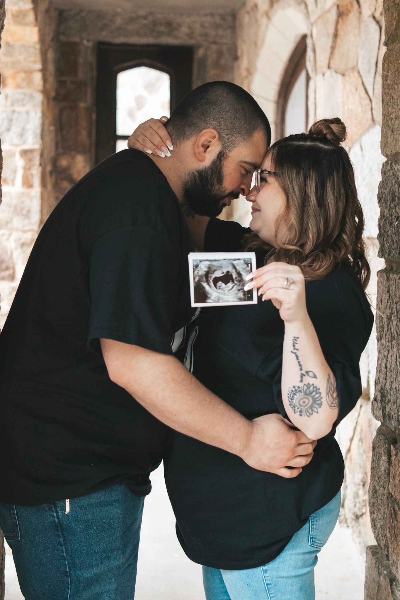 A romantic couple in black shirts and jeans embraces and kisses in a rustic brick hallway.