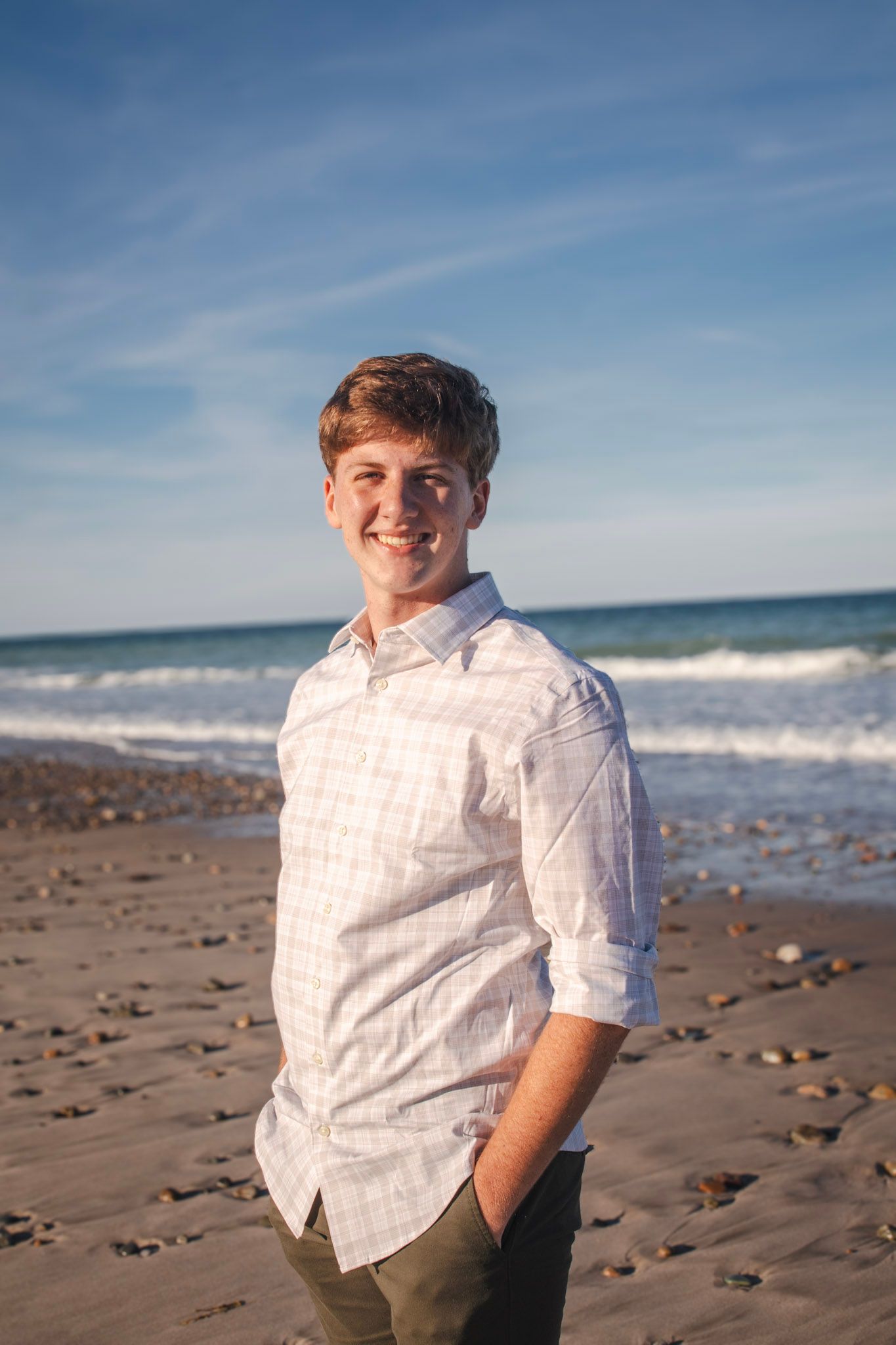 A person in a white shirt smiles while sitting on a sandy beach with ocean waves and blue sky in the background.