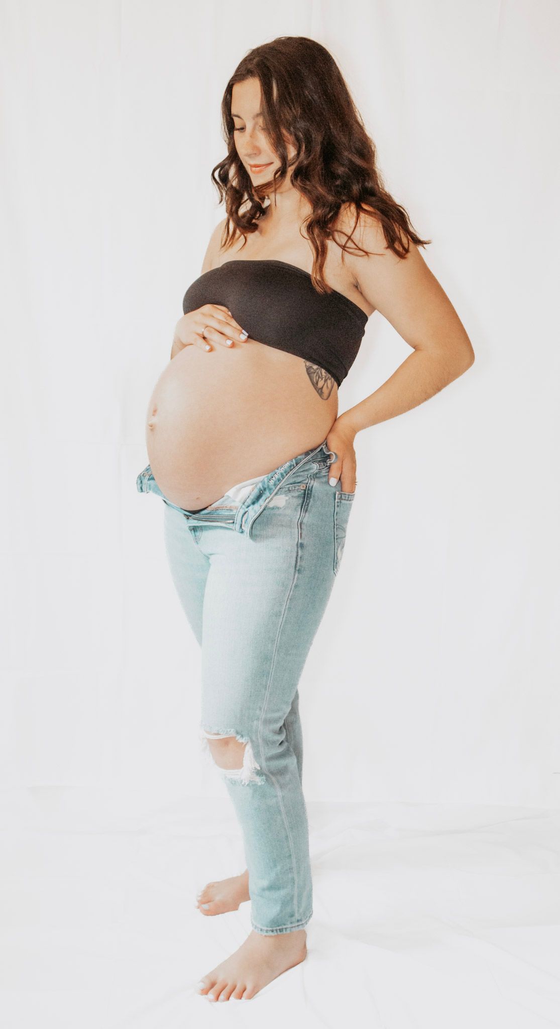 Woman in black top and light distressed jeans poses against white backdrop in artistic maternity photoshoot.