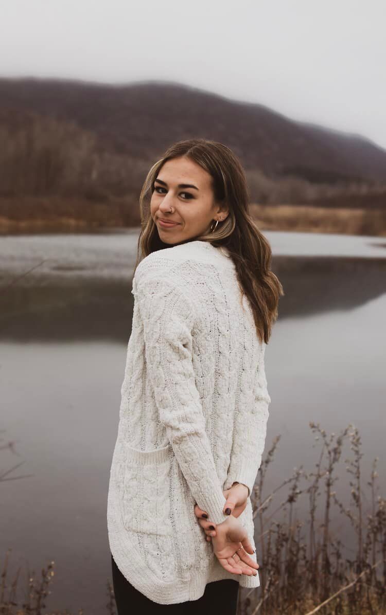 Person in white knit sweater poses against lake backdrop on cloudy day.