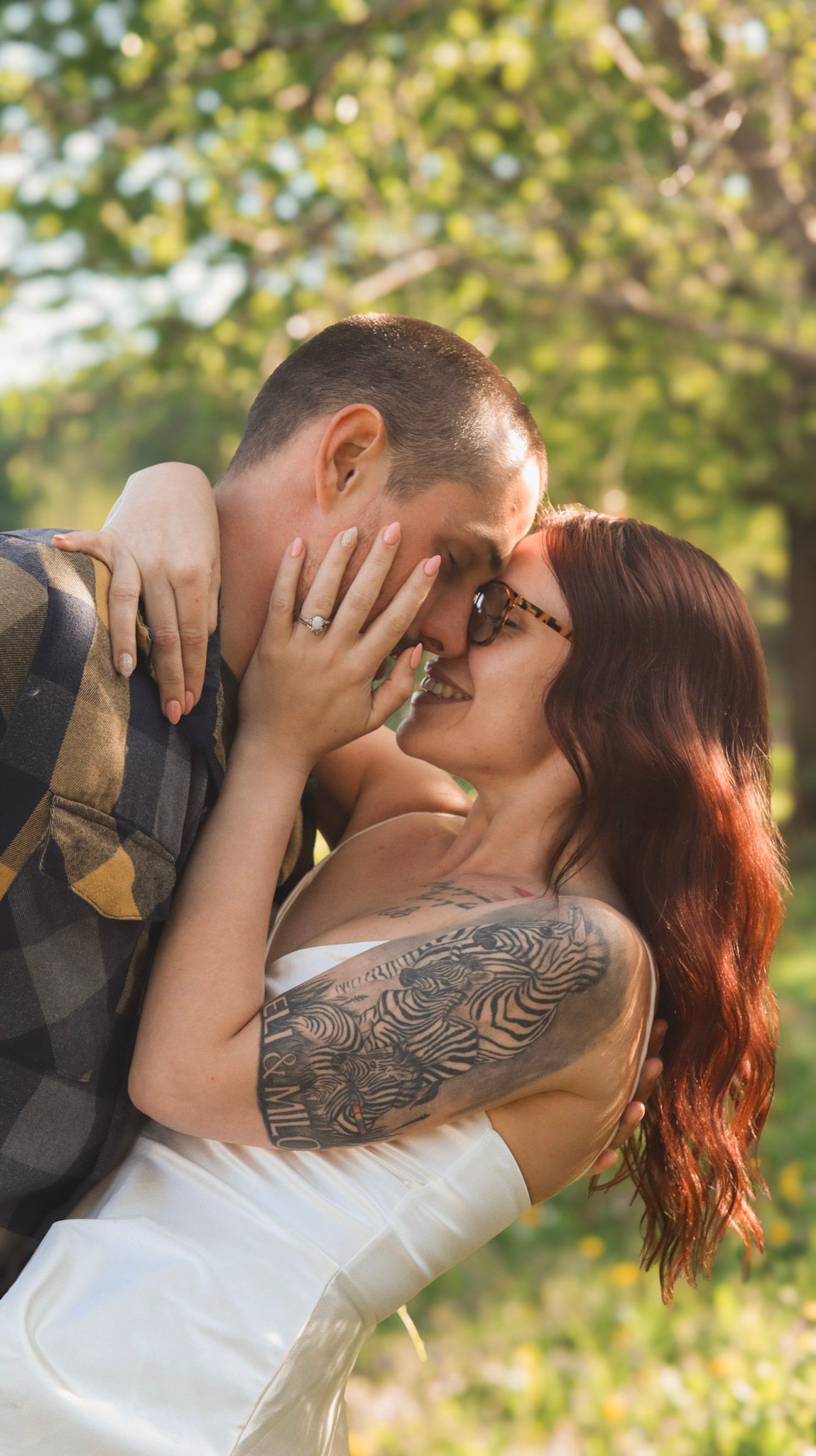 A romantic couple embraces and kisses in a sunlit outdoor setting with green foliage in the background.
