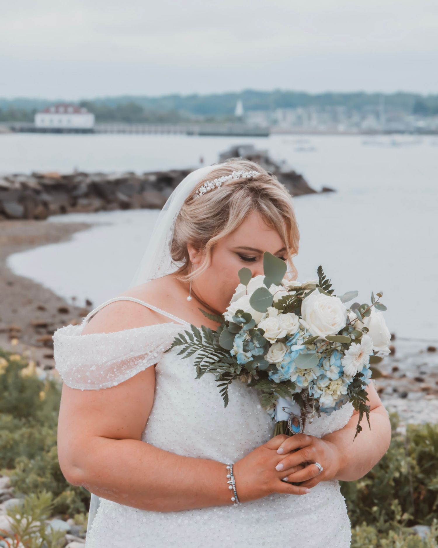 A bride in a white lace dress holds a bouquet of white and green flowers against a coastal landscape backdrop.