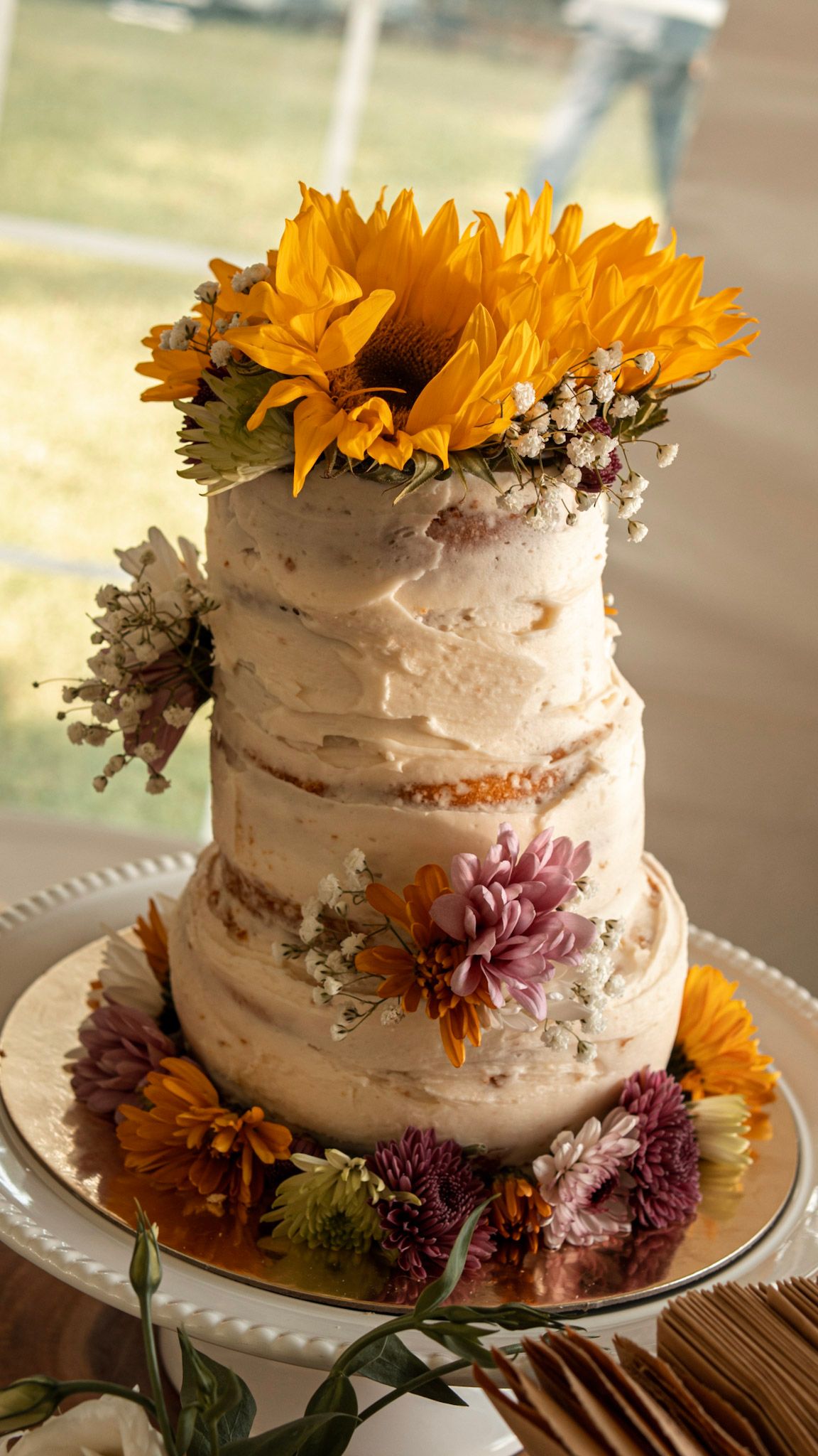 A semi-naked wedding cake decorated with sunflowers and purple flowers on a white cake stand.