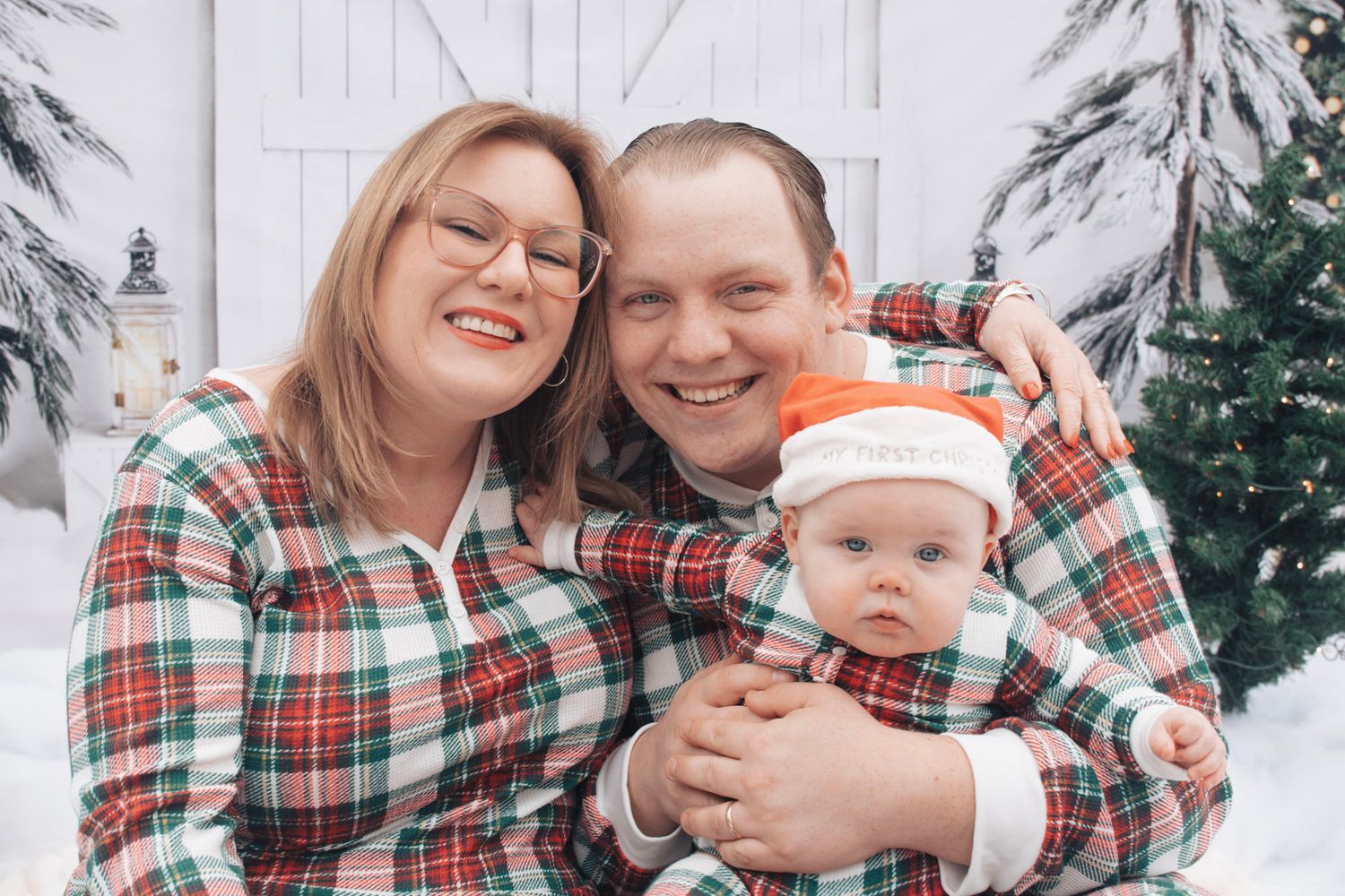 Family wearing matching plaid Christmas outfits posing in snow with white barn background.