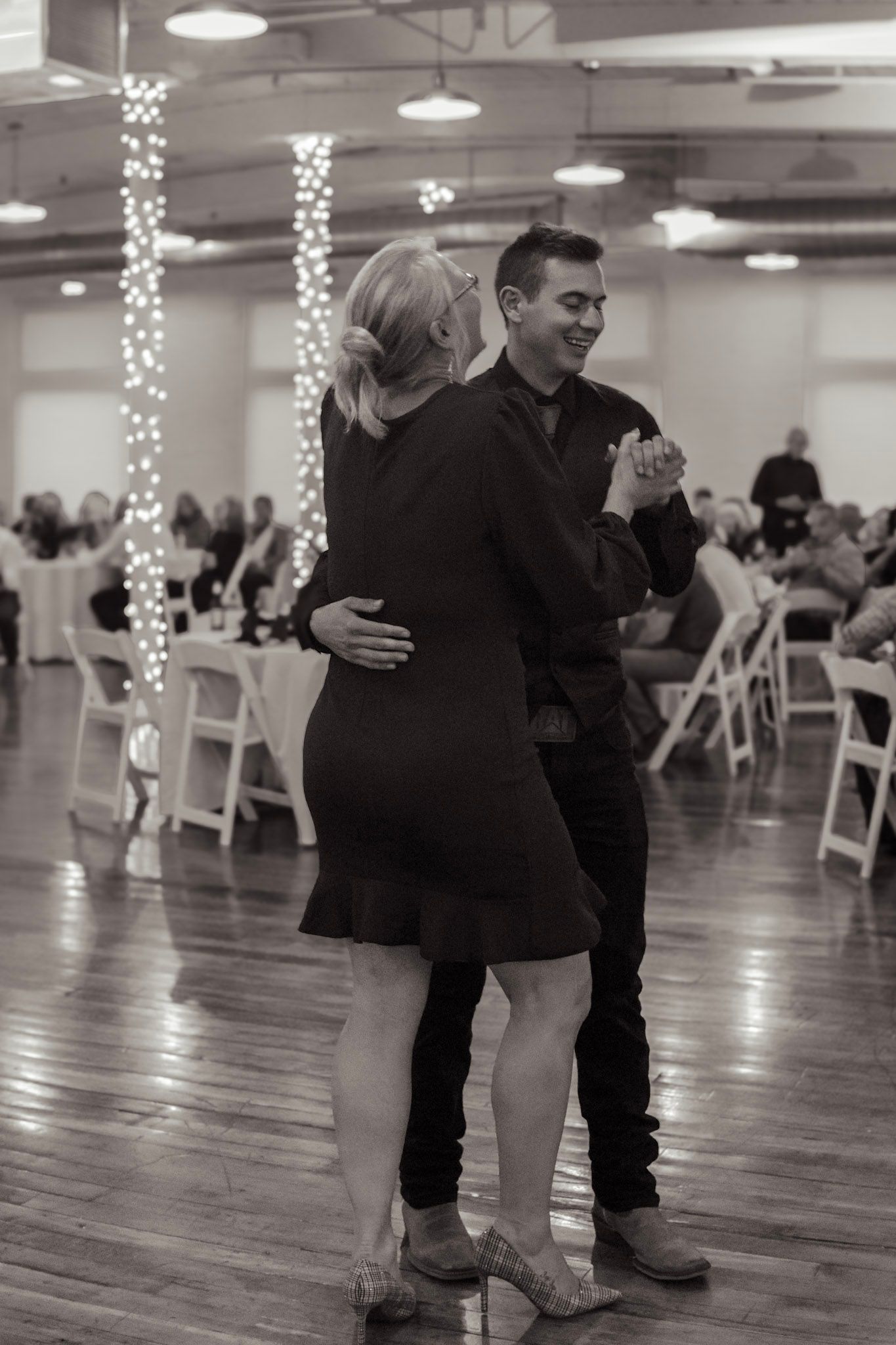 A couple slow dances together in a reception hall with string lights and white chairs visible in the background.