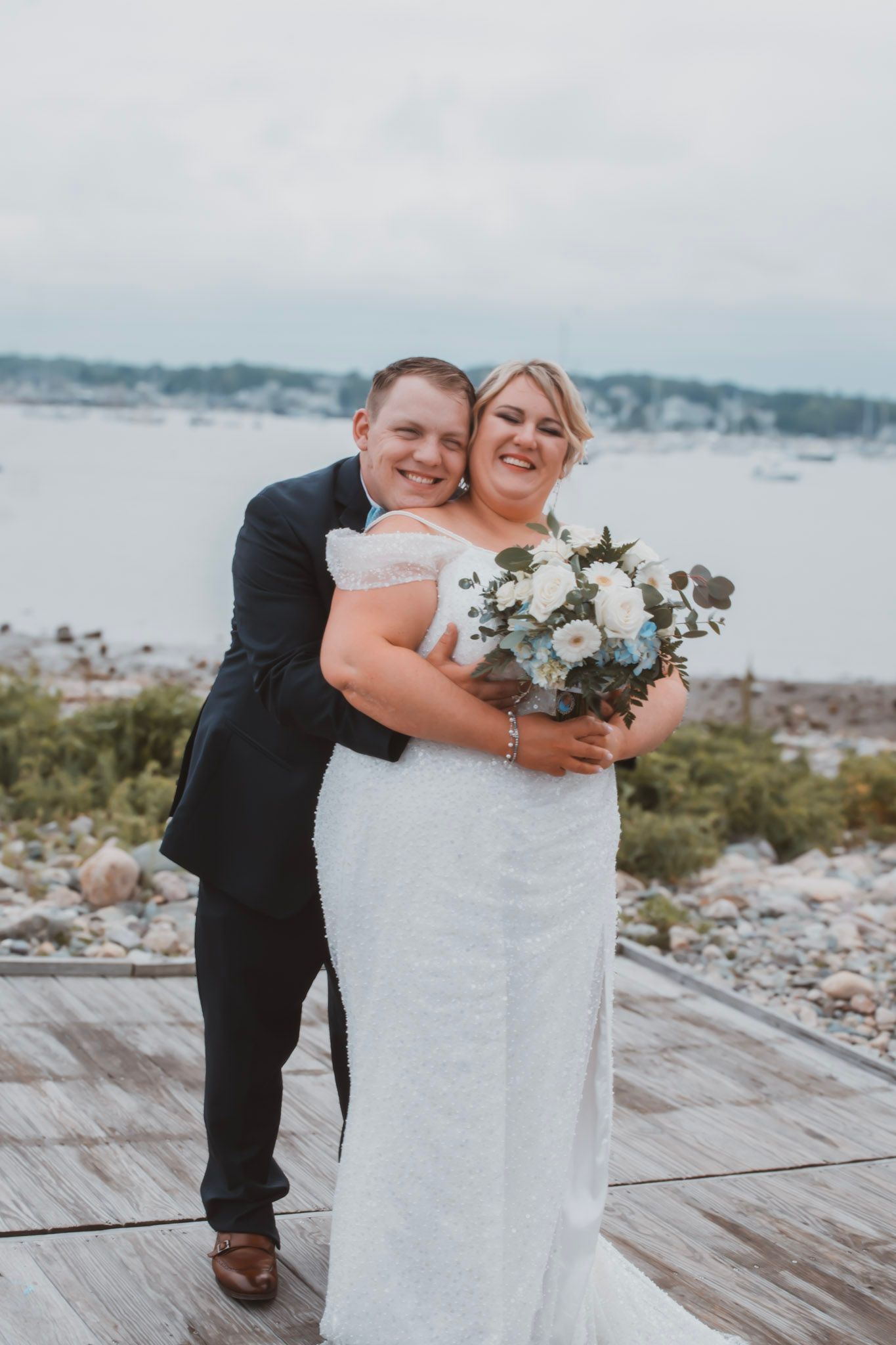 A joyful wedding couple sharing a sweet moment on a waterfront deck with a beautiful bouquet of white flowers.