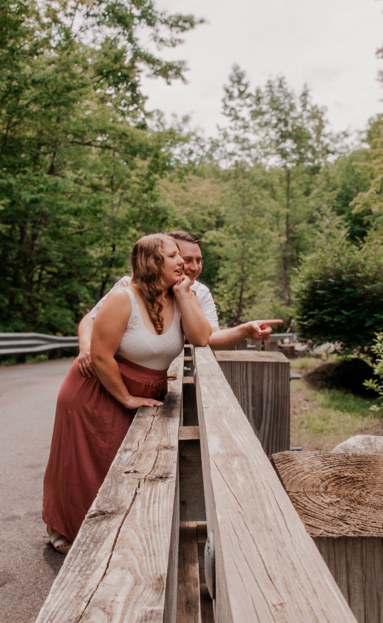 Two people in wedding attire share a tender moment while leaning over a wooden railing in a natural outdoor setting.