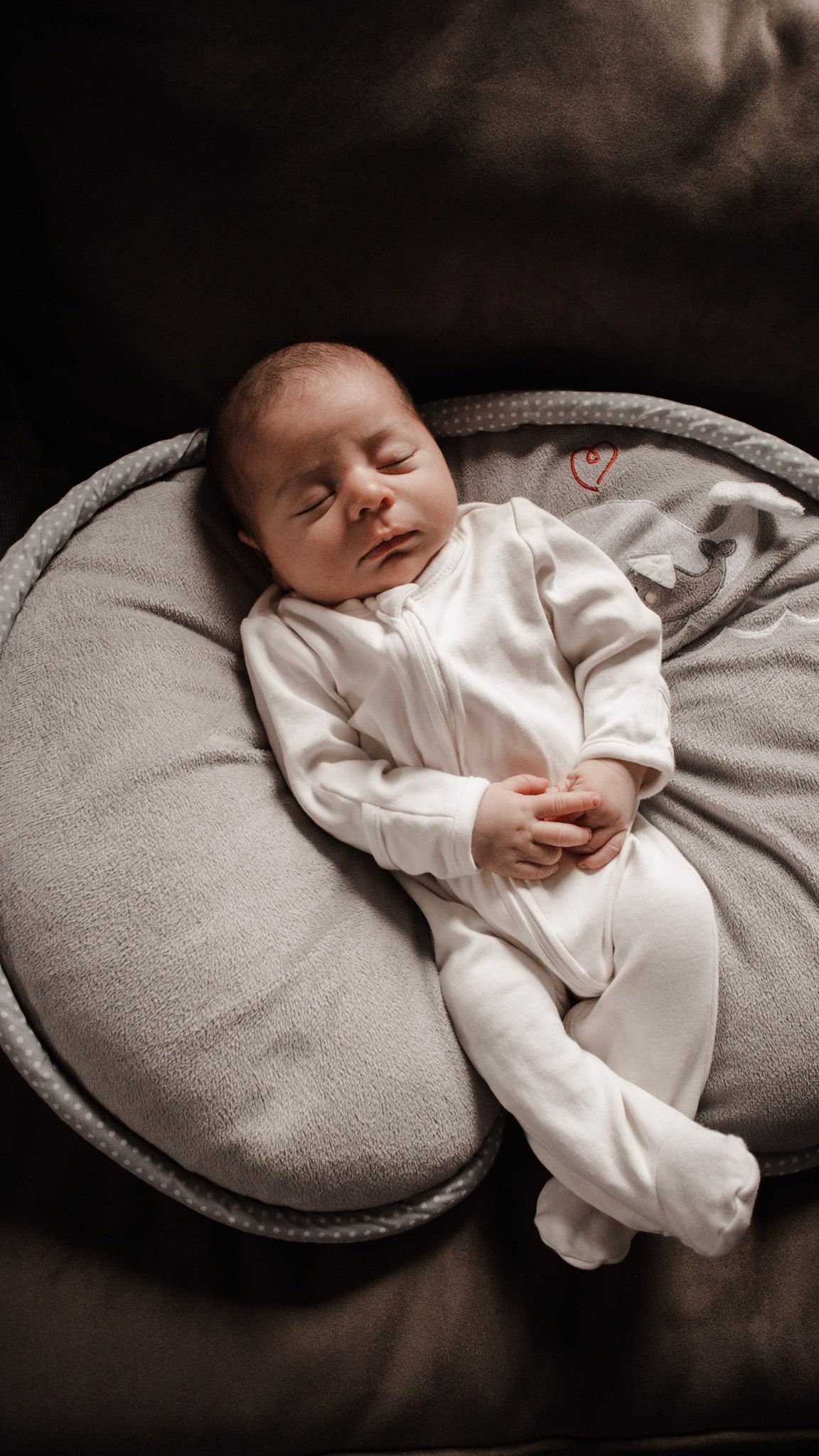 A sleeping newborn baby in white clothing rests peacefully on a curved gray nursing pillow.