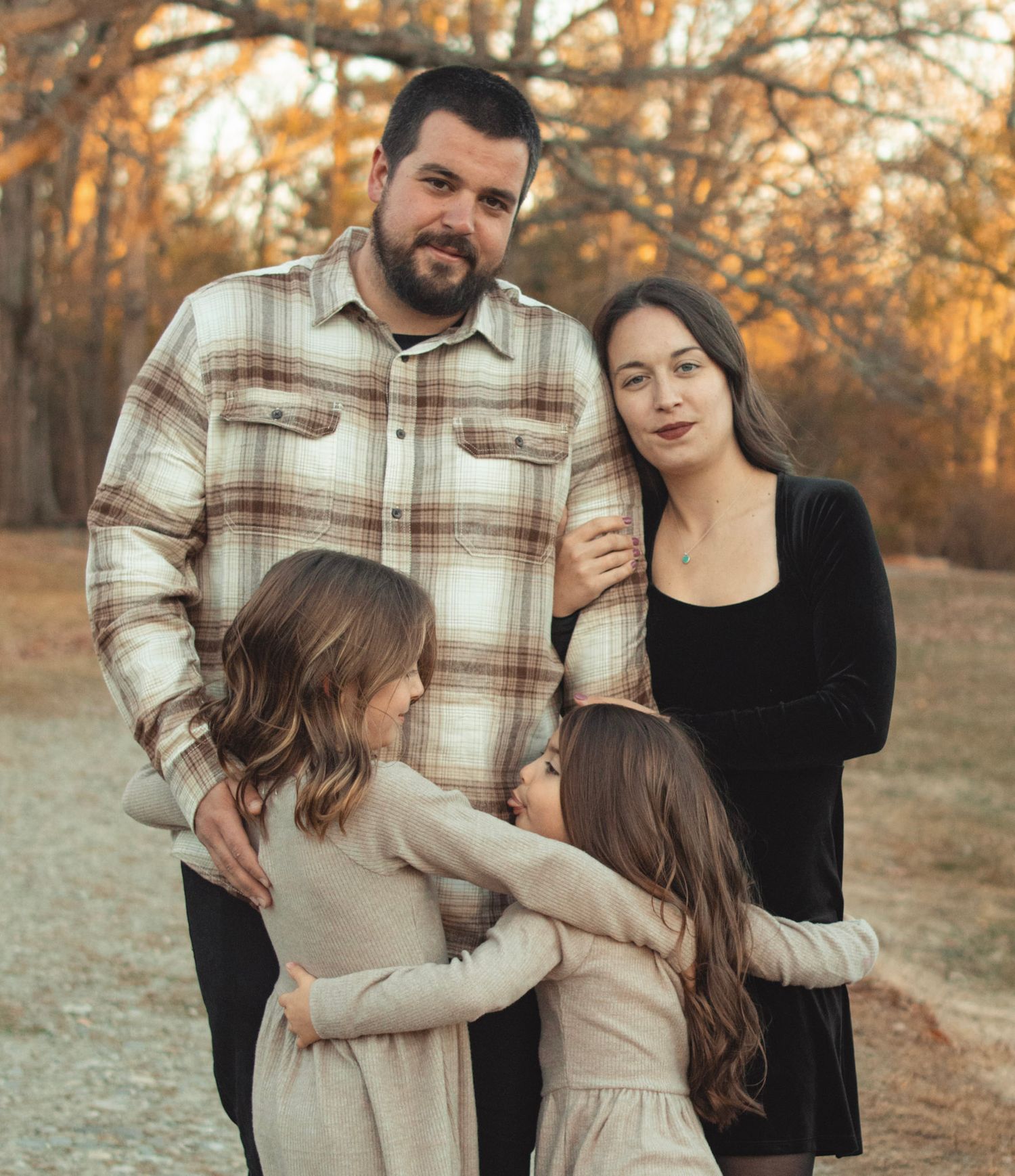 A family in coordinating autumn outfits embraces during a golden hour photoshoot in a rustic outdoor setting.