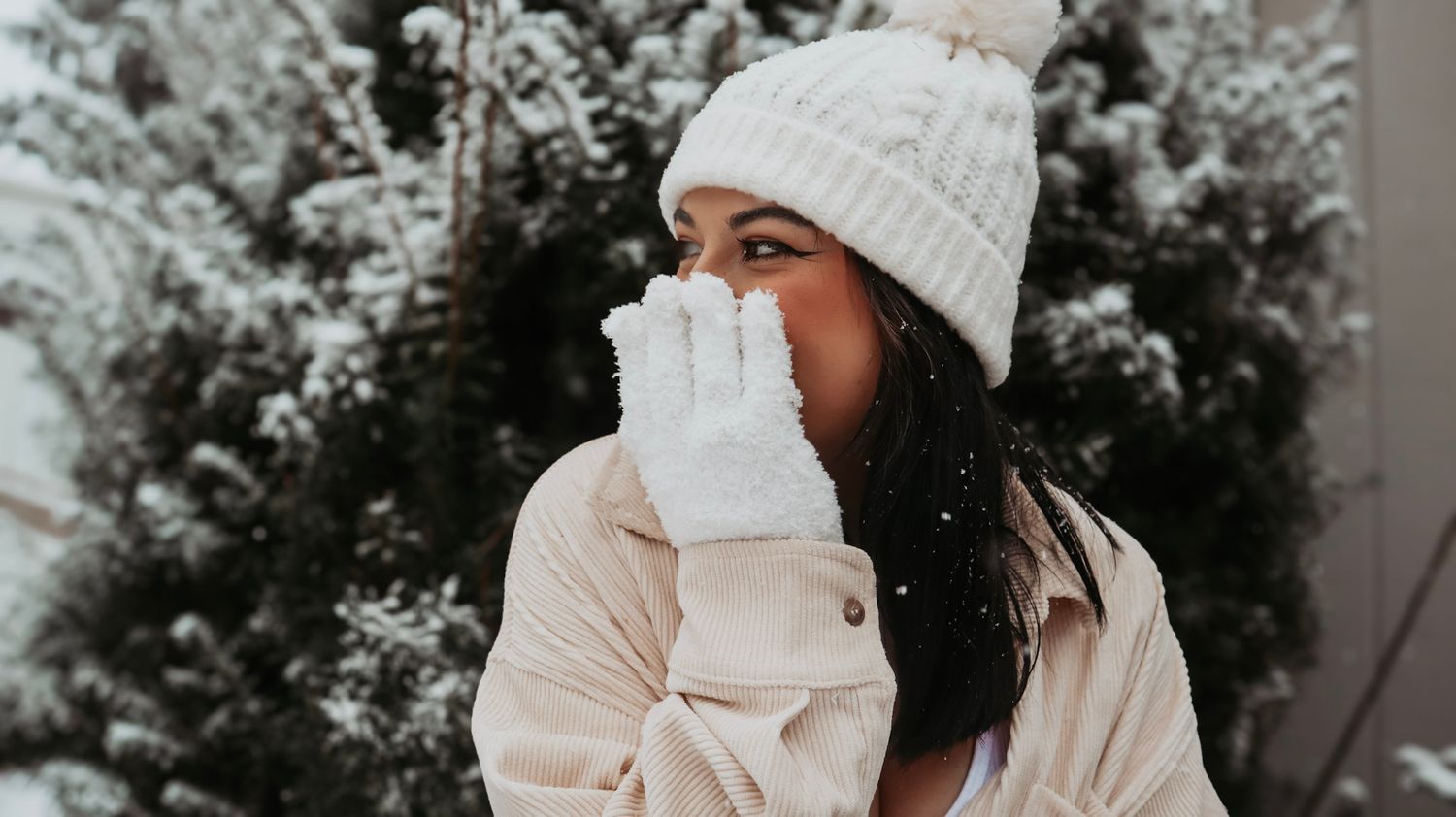 Person wearing a white knit beanie and mittens stands against a snowy winter backdrop with evergreen trees.