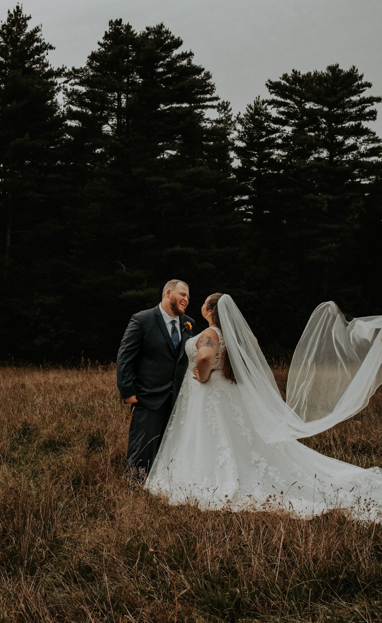 A couple in wedding attire share an intimate moment in a field with tall pine trees silhouetted against the sky at dusk.