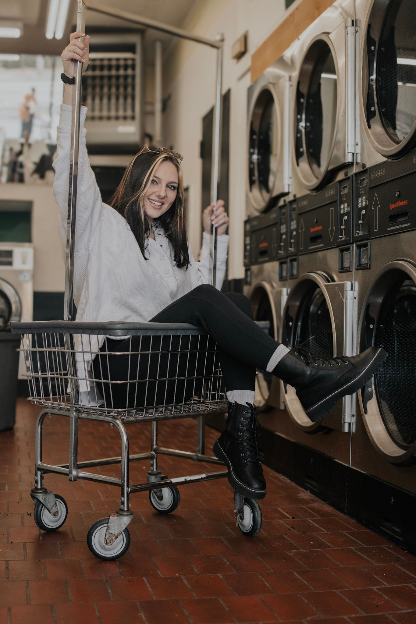 Person having fun riding in laundry cart while doing laundry at laundromat with washing machines in background.