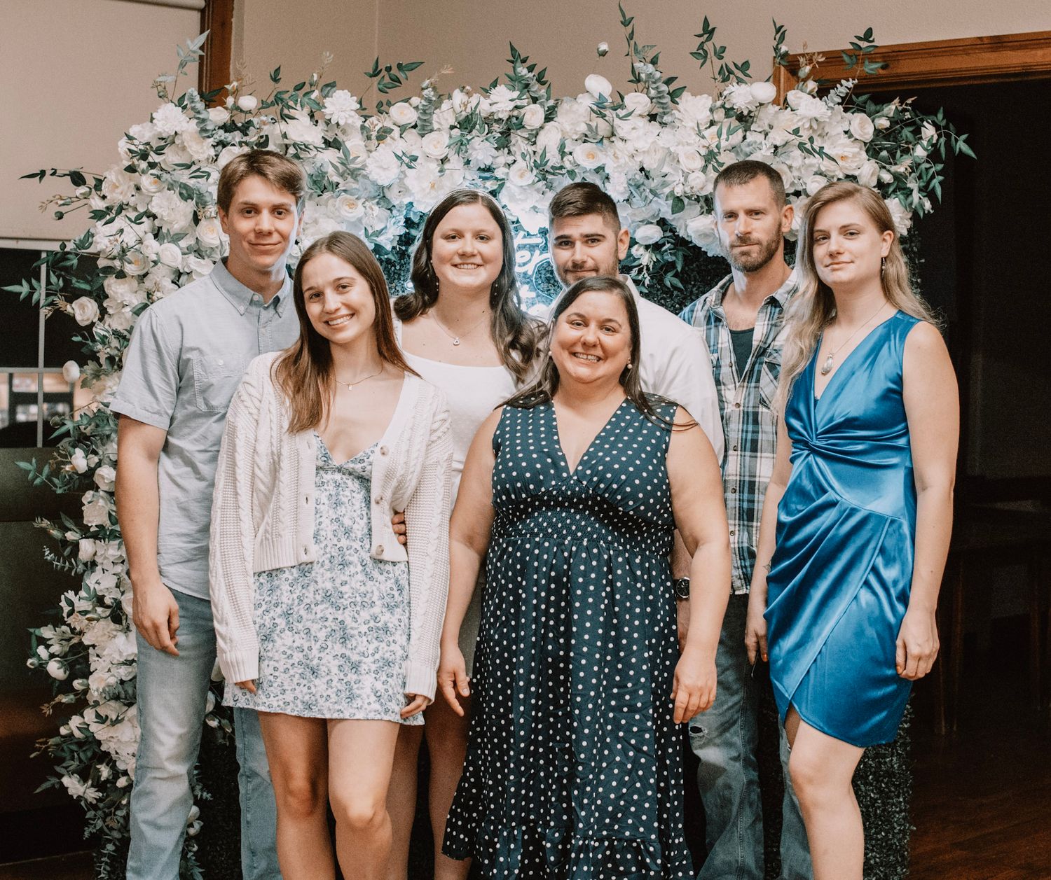 Group photo against a floral backdrop with people wearing coordinated blue and white outfits at a special event.