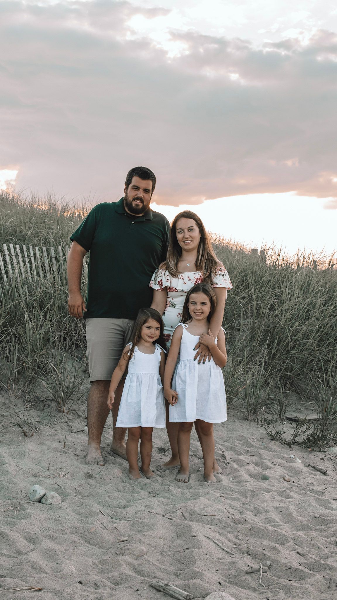A family poses together on a sandy beach at sunset with tall grass and a wooden fence in the background.