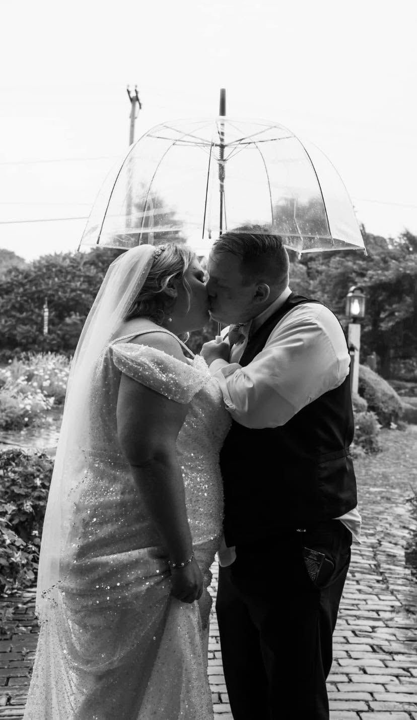 Black and white wedding photo of a couple kissing under a clear umbrella on a brick pathway.