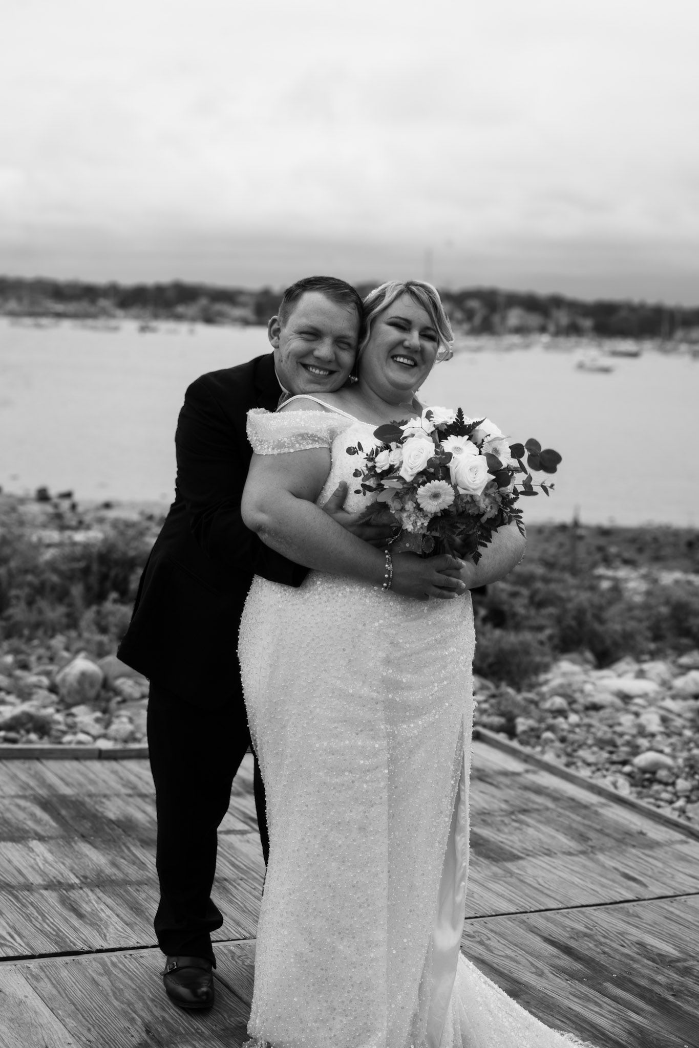 A romantic wedding couple embraces on a wooden deck overlooking a scenic harbor view on their special day.