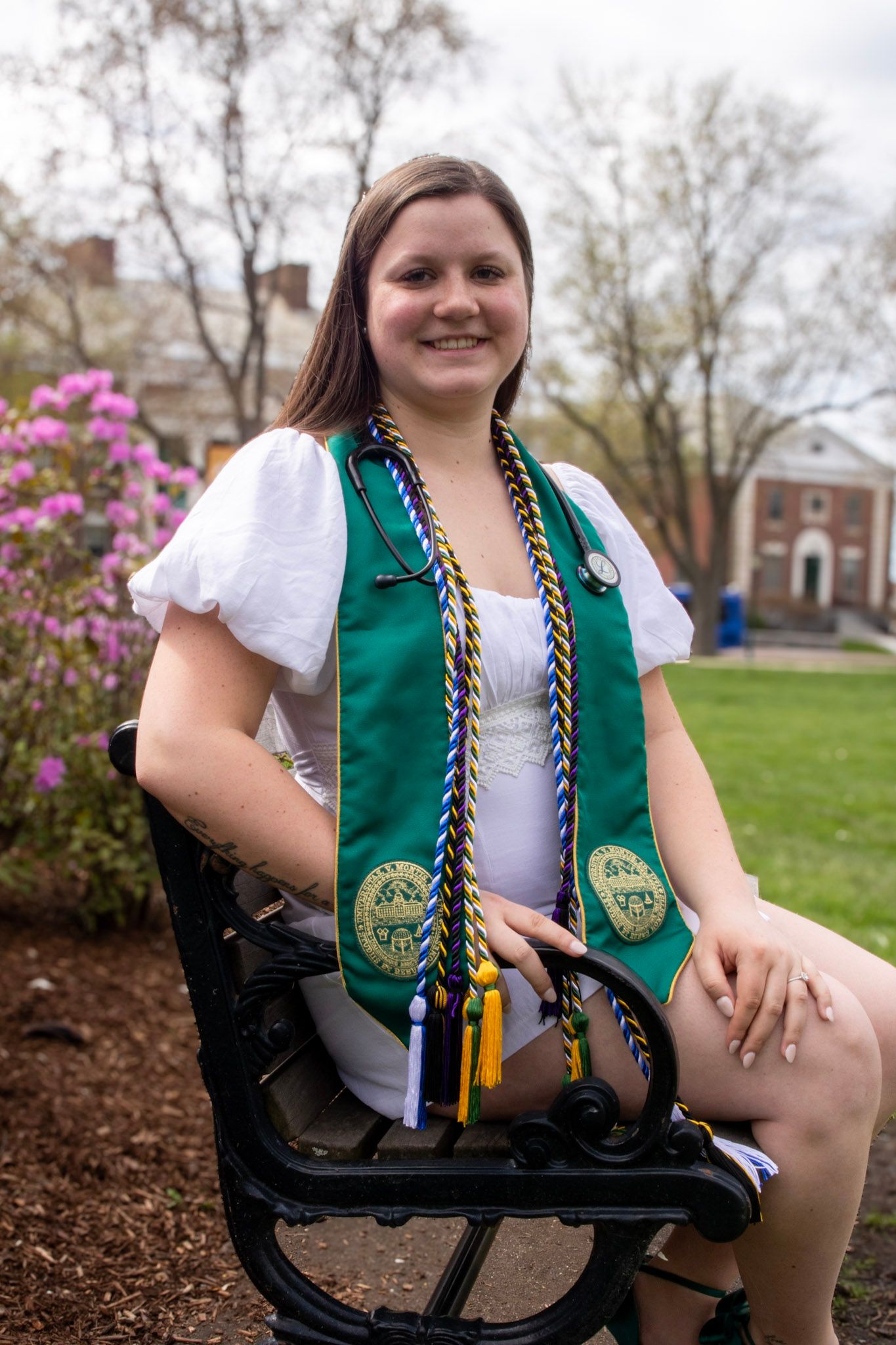 A graduate in a green and gold honor stole sits on a bench surrounded by spring blooms and trees on a college campus.