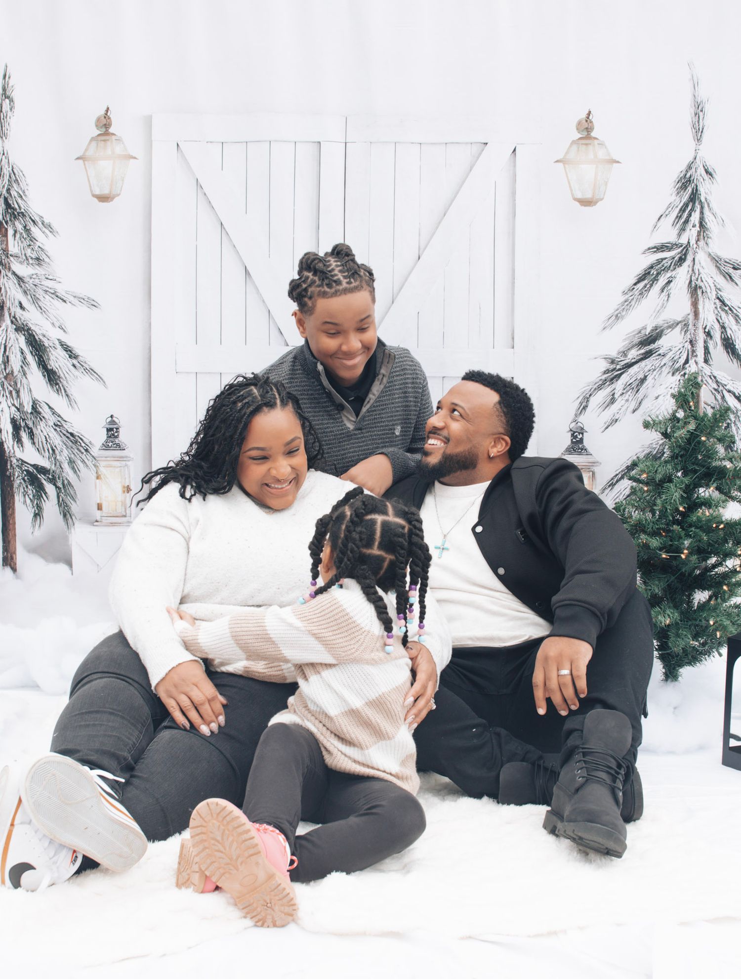 A couple playfully interacts while sitting in the snow against a white fence with evergreen trees in the background.