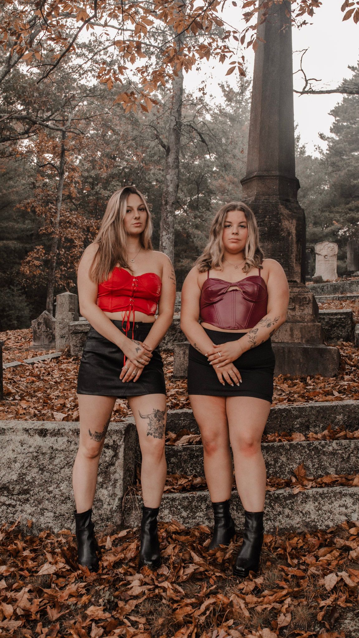 Two figures in red tops and black skirts pose among autumn leaves in a moody cemetery setting with stone monuments.