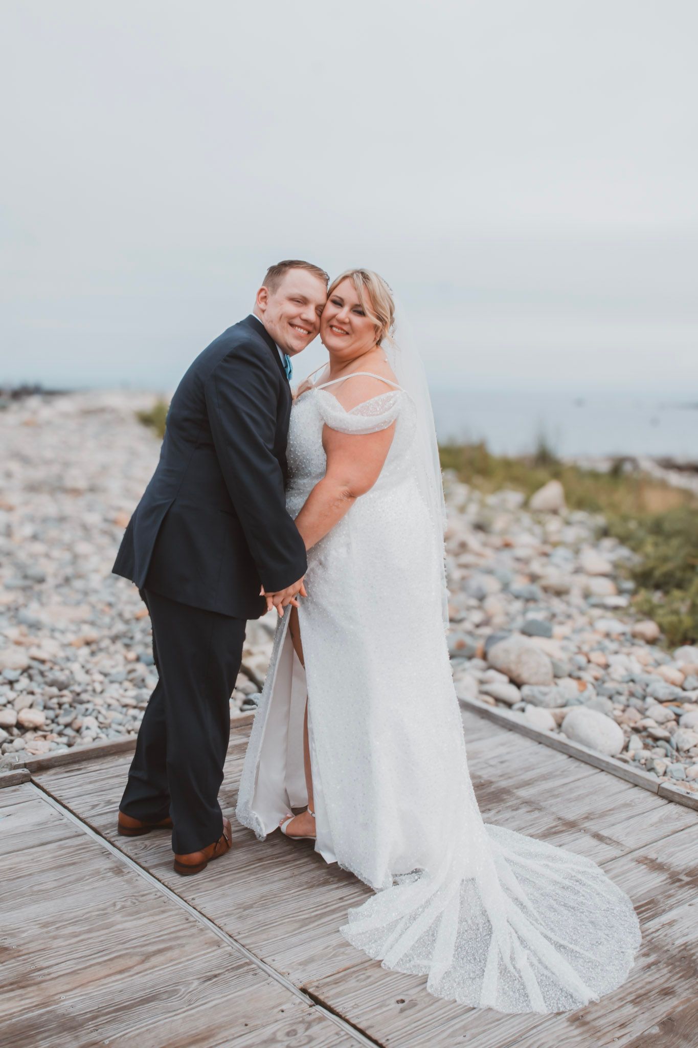 A couple shares a romantic moment on a wooden boardwalk by a rocky beach during their wedding photoshoot.