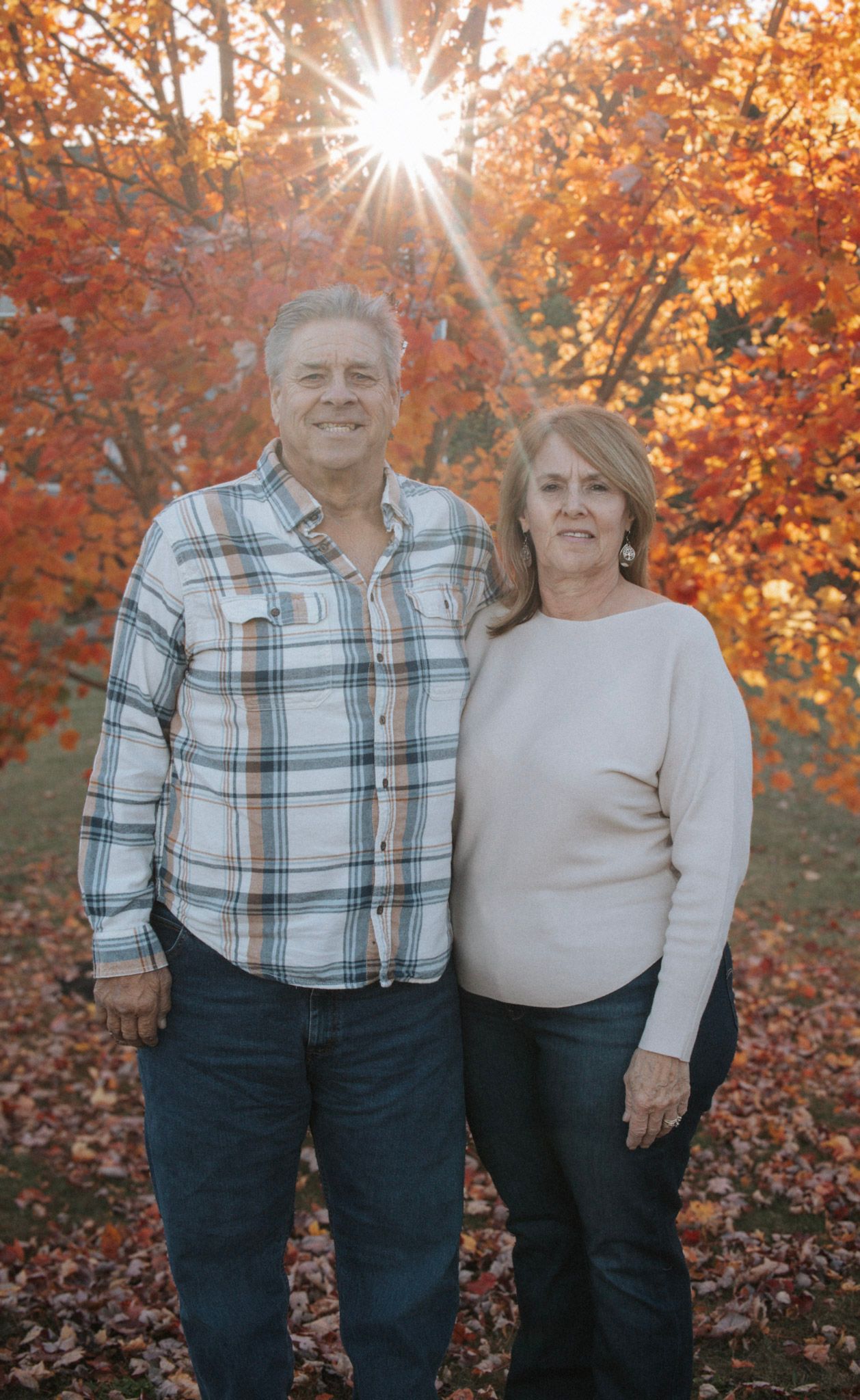 Autumn portrait of a couple standing together among vibrant orange fall foliage with sunburst behind them.