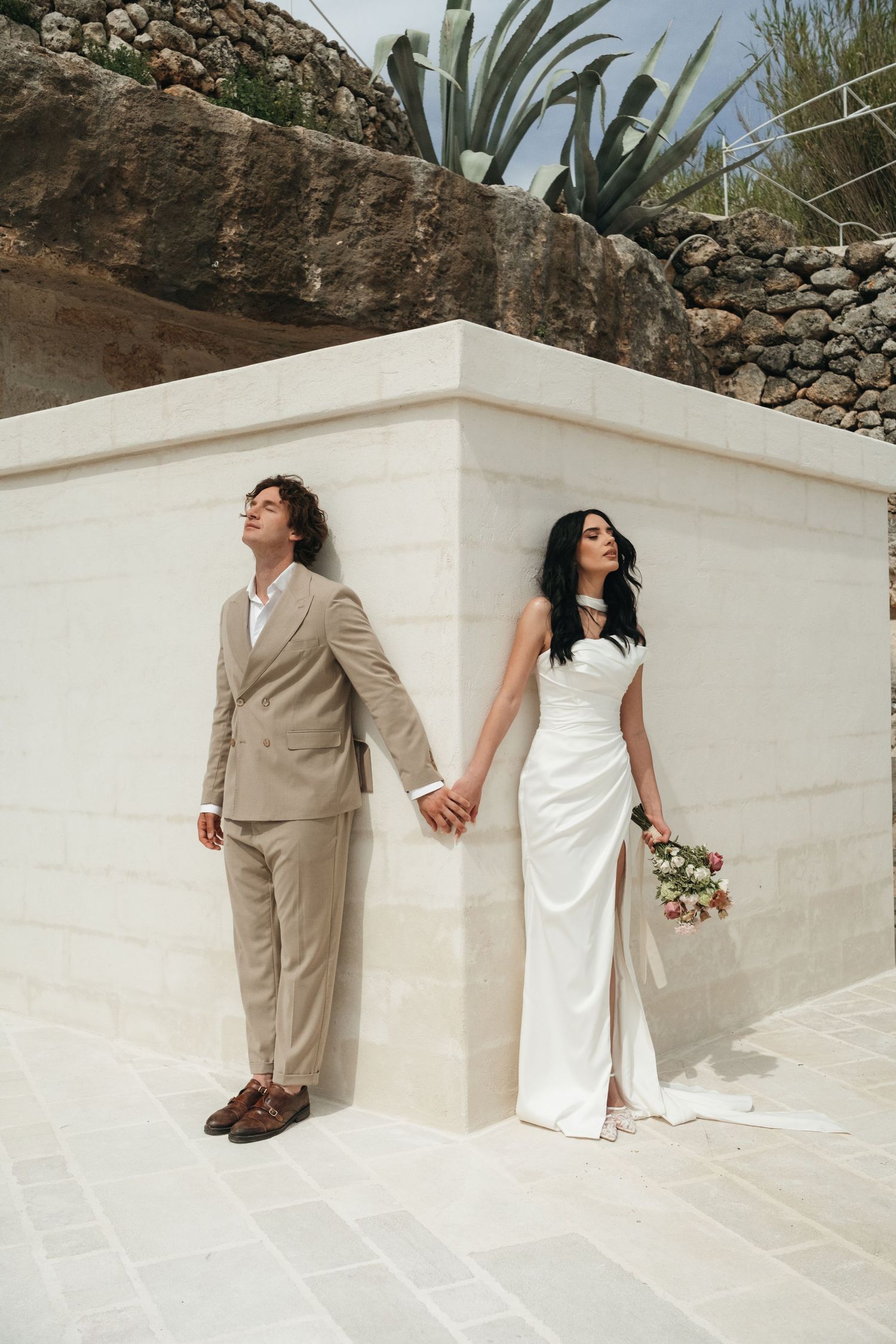 Bride in white gown and groom in beige suit hold hands leaning against a stone wall outdoors.
