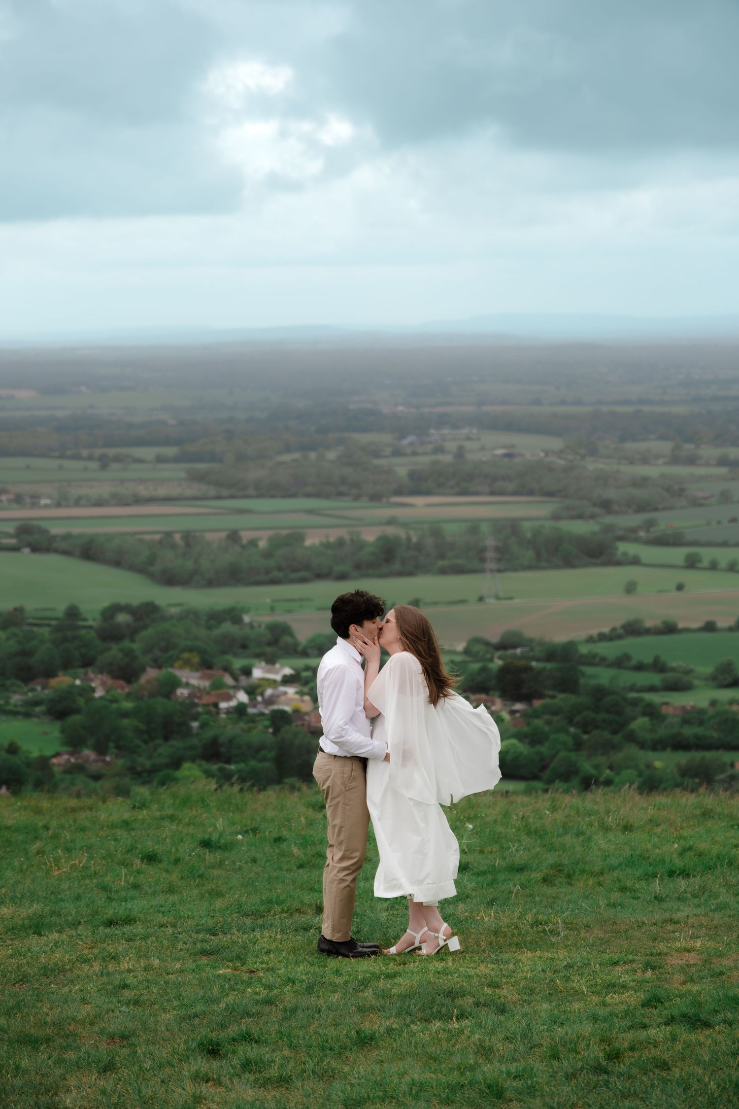 Couple sharing a romantic kiss on a hilltop in white wedding attire with scenic green countryside below.